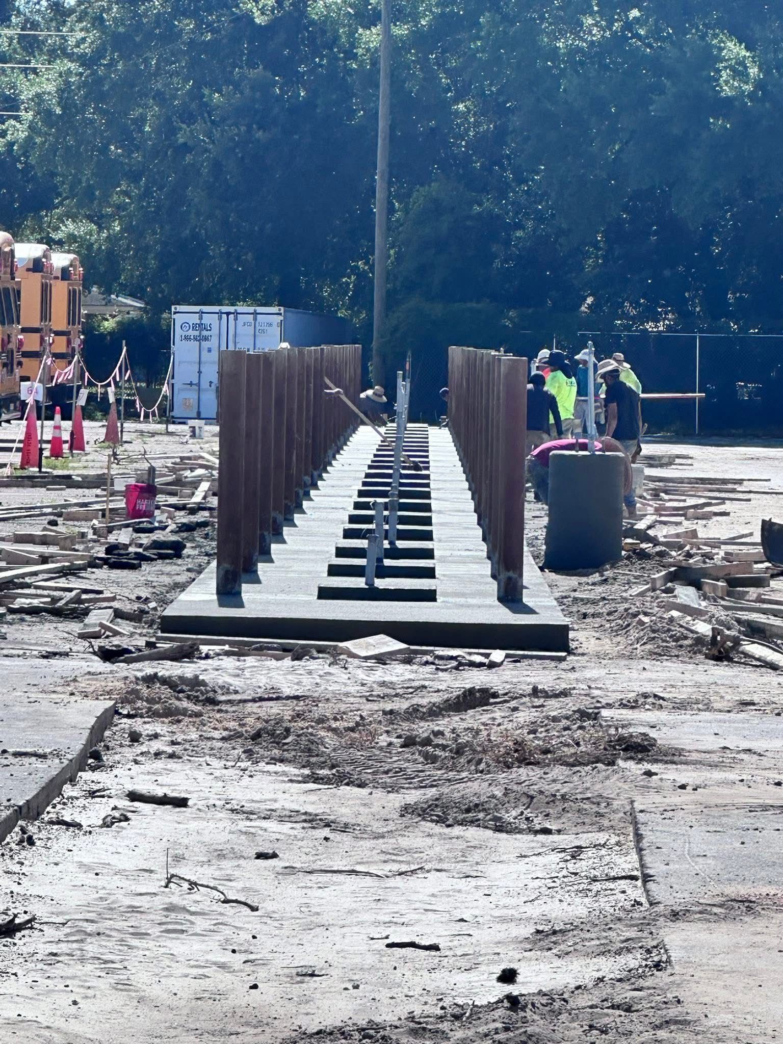 Wooden boardwalk ramp under construction, with railings, on a muddy site surrounded by trees.