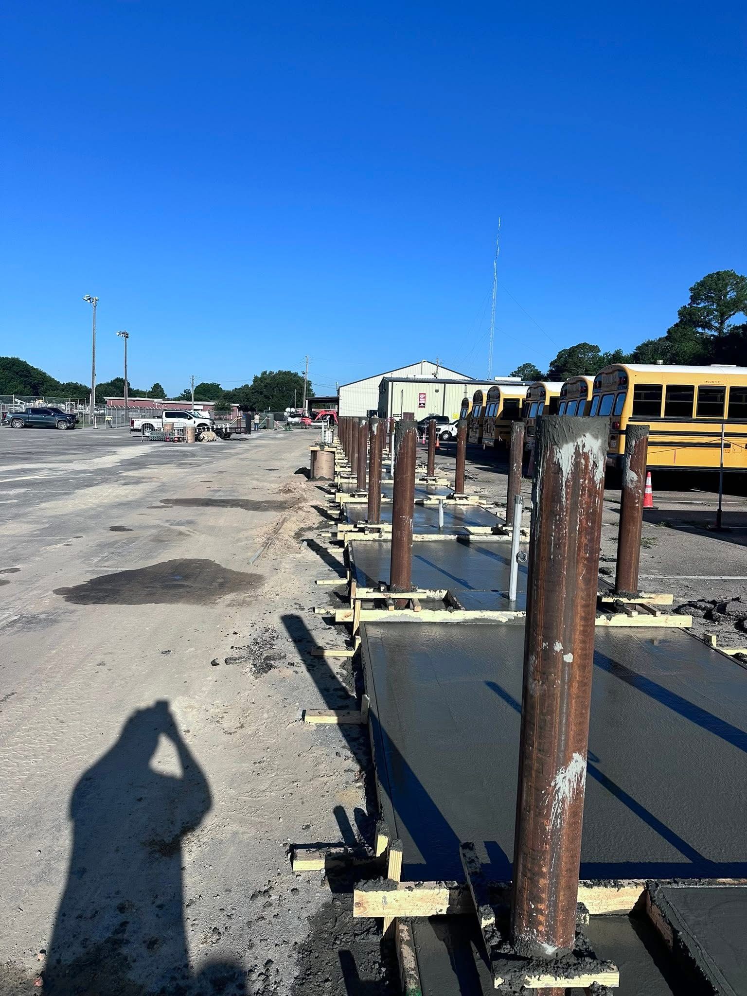 Construction site with rusted metal posts, asphalt, and a yellow school bus in the background under a clear blue sky