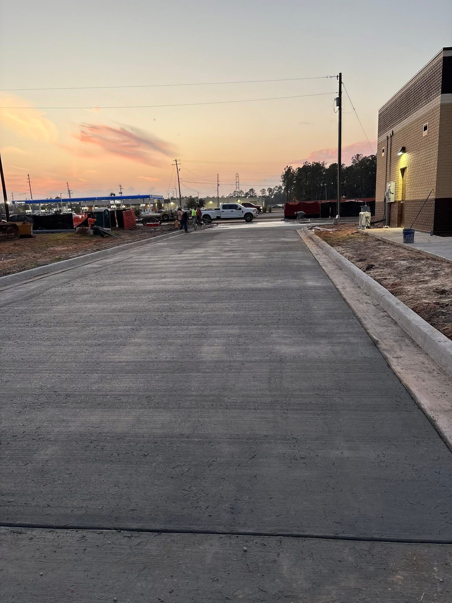 Wide concrete driveway beside a brick building at sunset, with parked cars and utility poles in the distance.