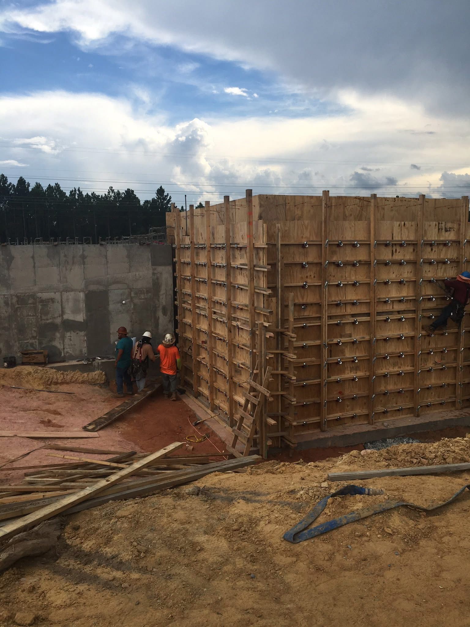 Workers pour concrete around a large wooden form at a construction site under a cloudy sky.
