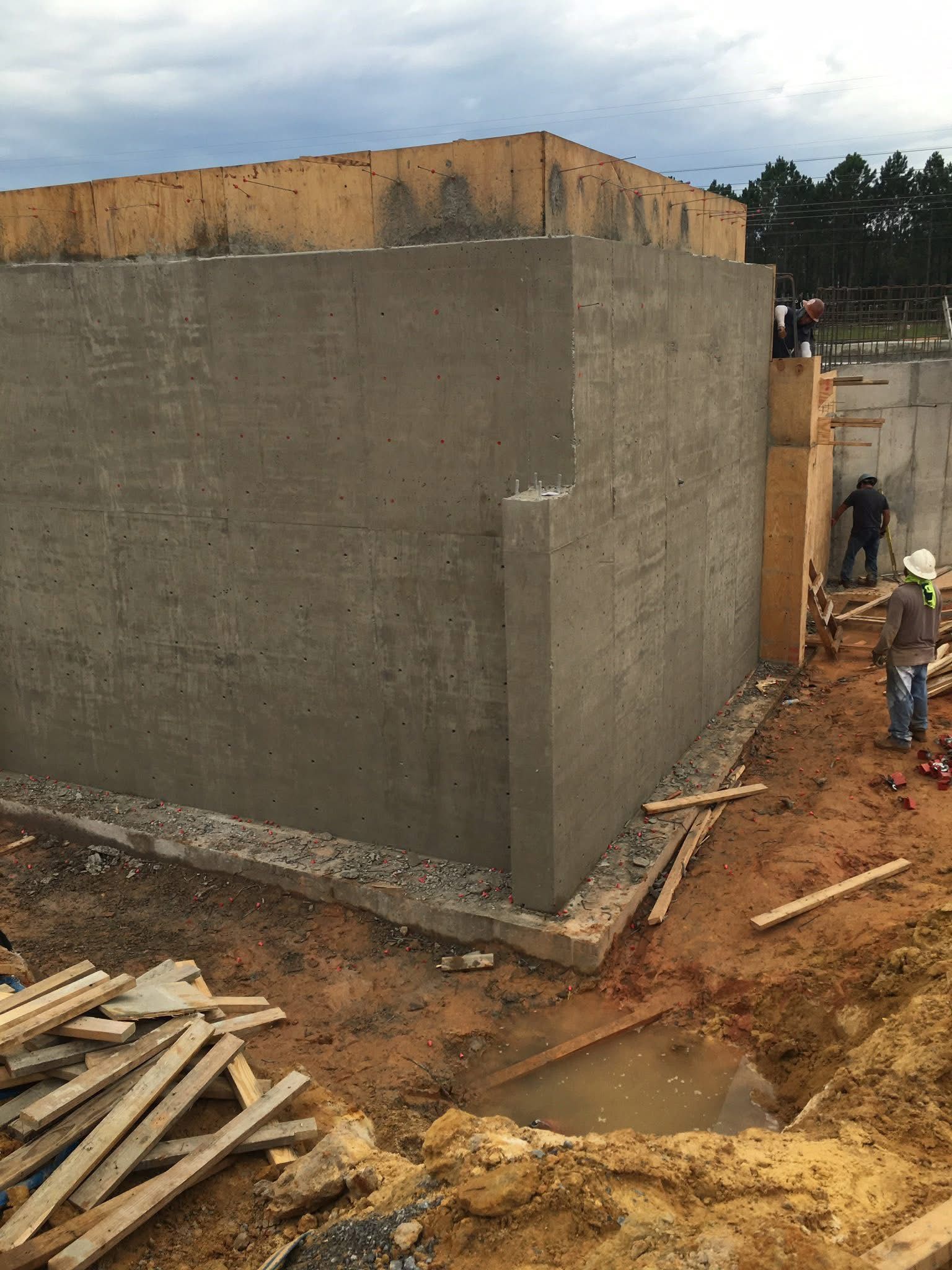 Large concrete building under construction with workers and lumber on muddy ground.