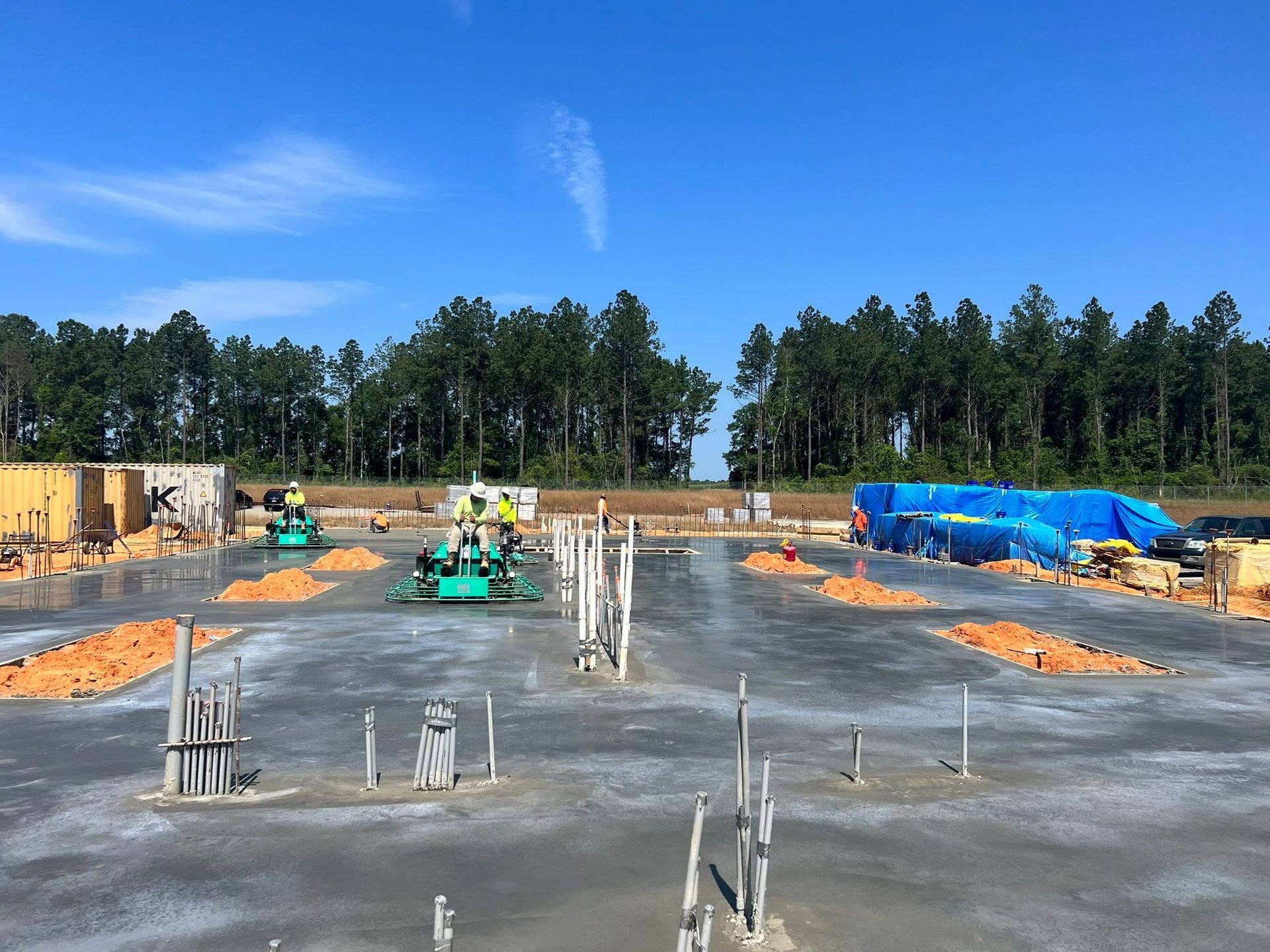 Construction site with concrete pads, equipment, blue tarps, and trees under a clear sky