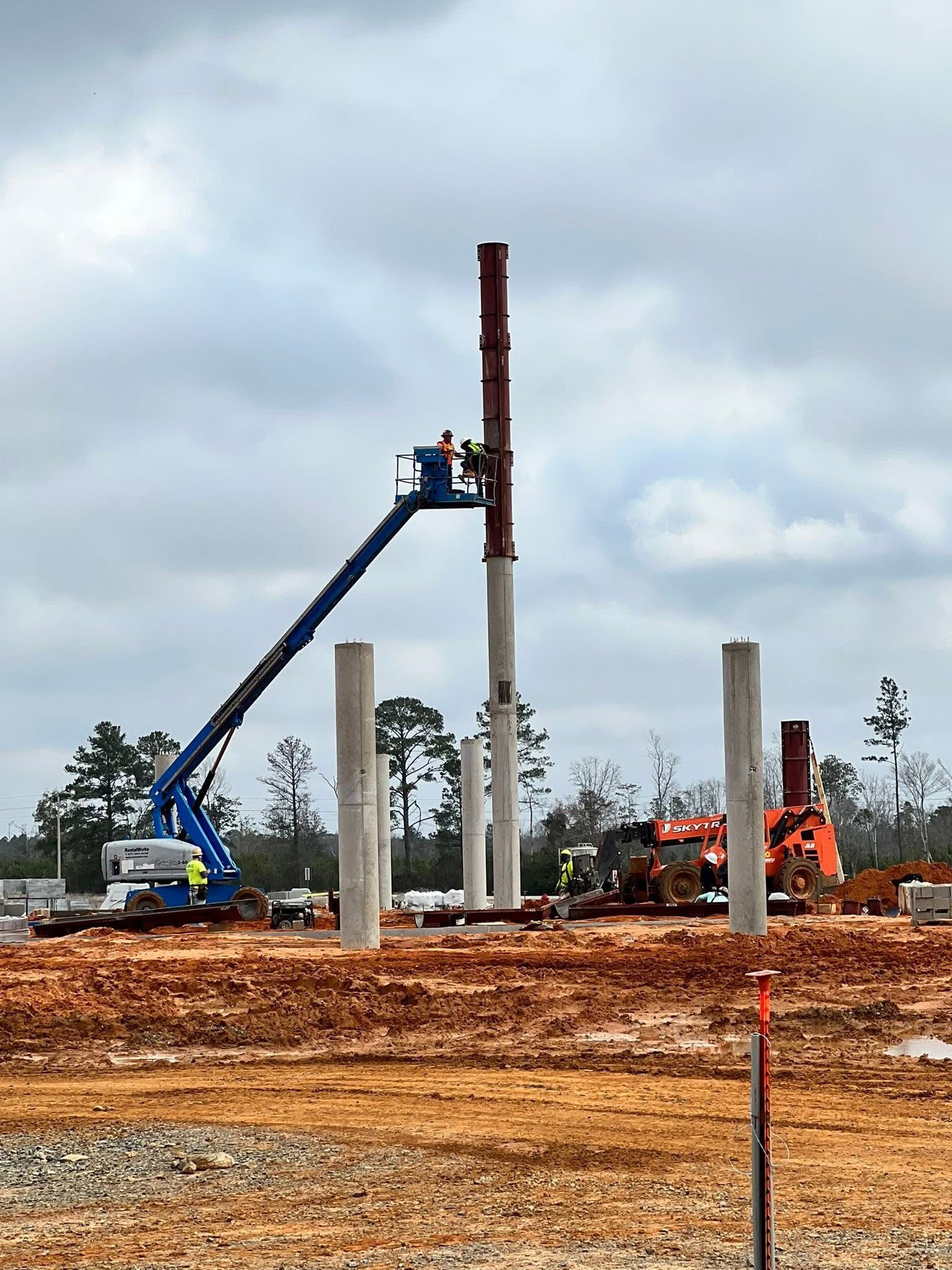 Construction site with tall concrete pillars, a blue boom lift, and orange excavators on muddy ground.