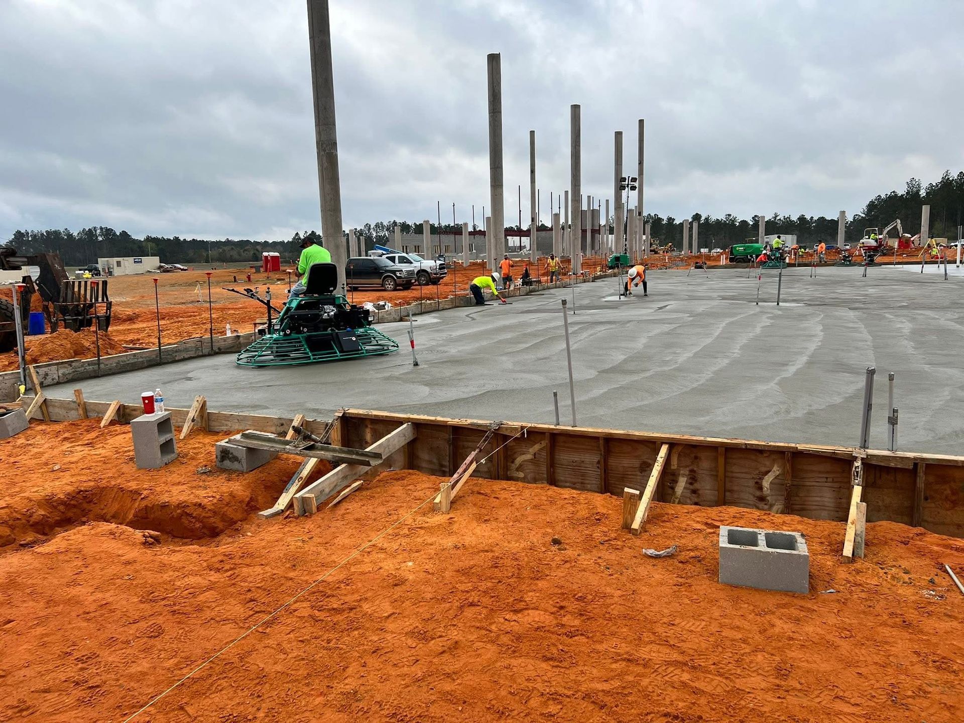 Construction site with concrete slab, vertical steel posts, and orange dirt, with workers in the distance