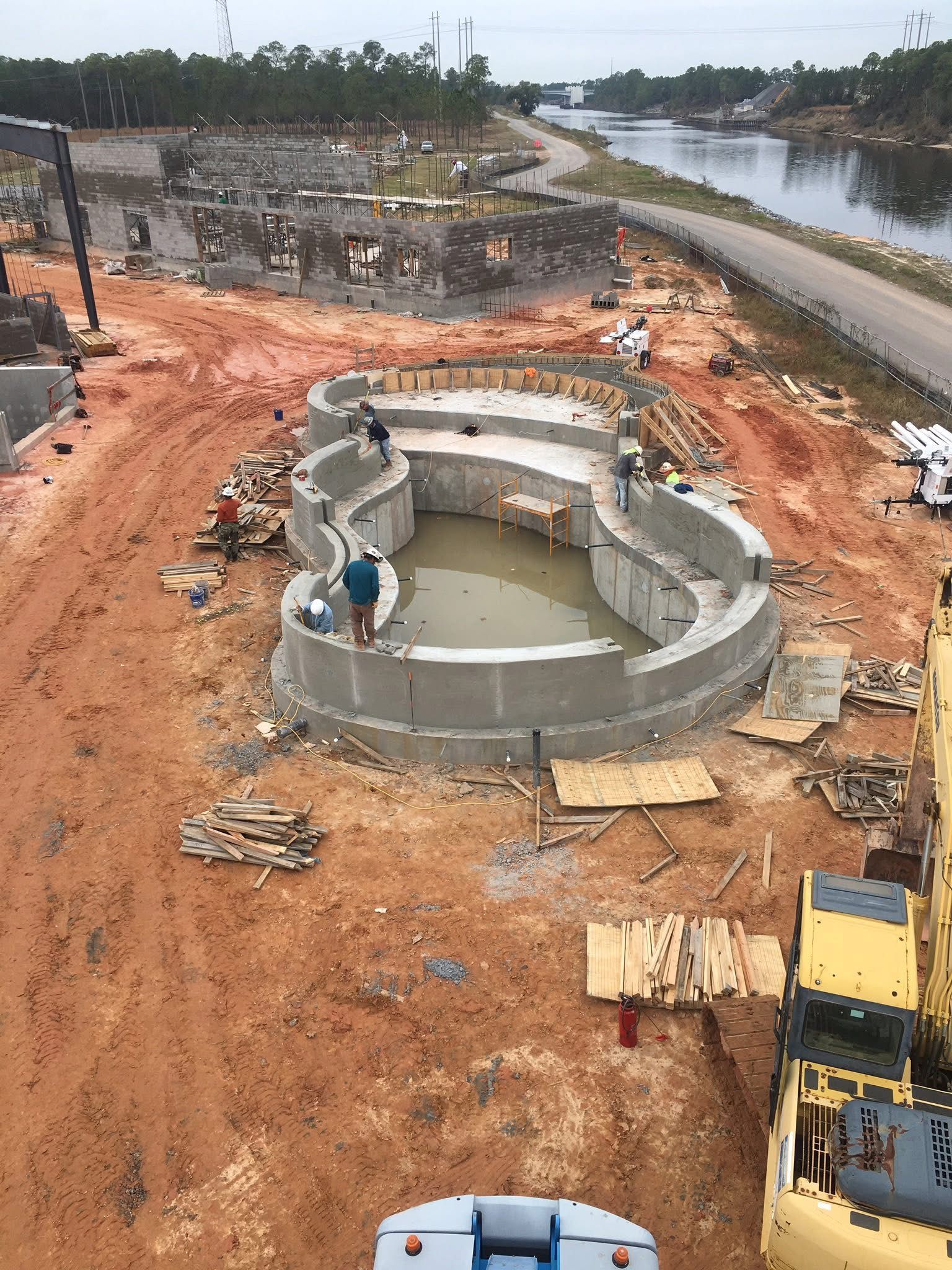 Construction site with a circular concrete structure, workers, earthmoving equipment, and a canal nearby