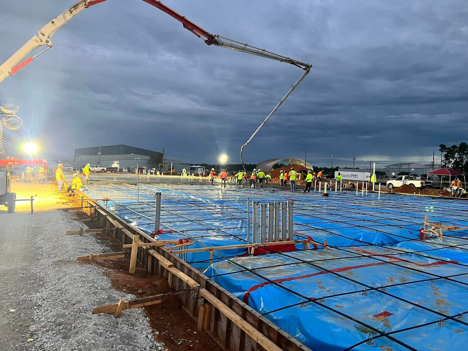 Construction site at dusk with concrete pump pouring onto blue-covered foundation and workers nearby