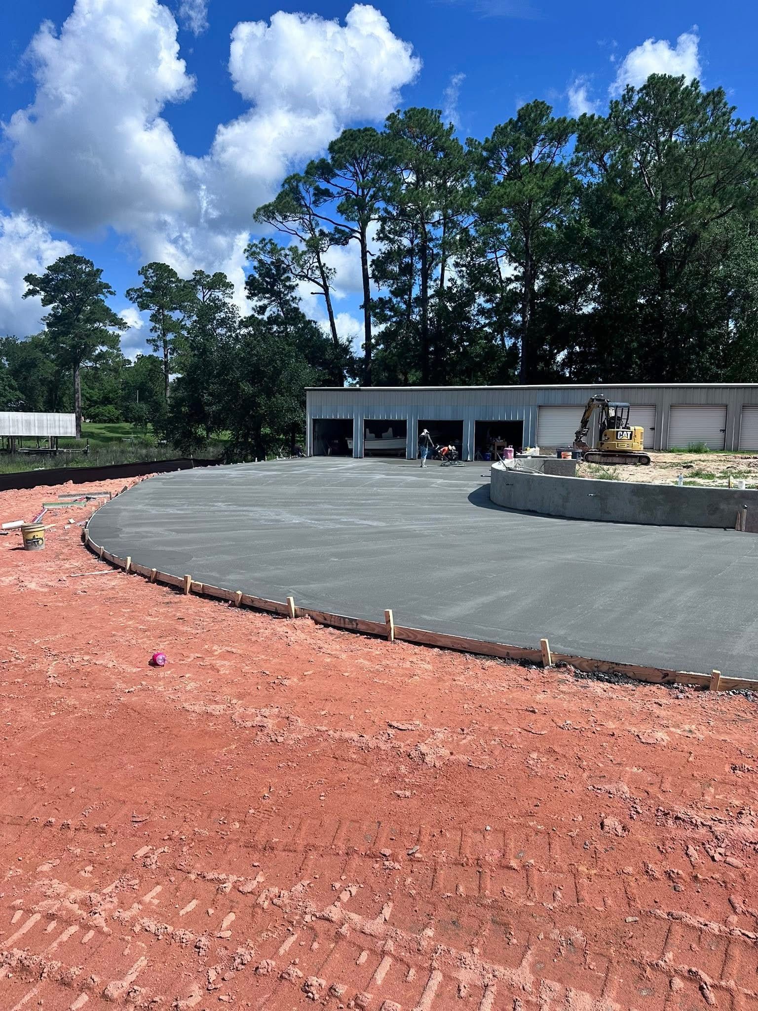 Curved asphalt path beside a building under construction, with red dirt foreground and trees under a blue sky.