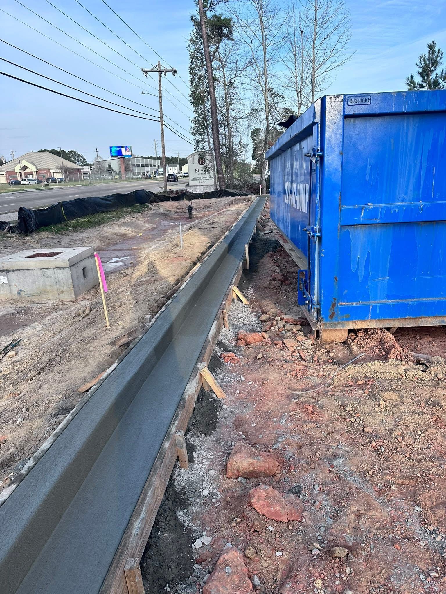 Blue utility shed beside a roadside drainage ditch and metal guardrail on a bare, muddy lot