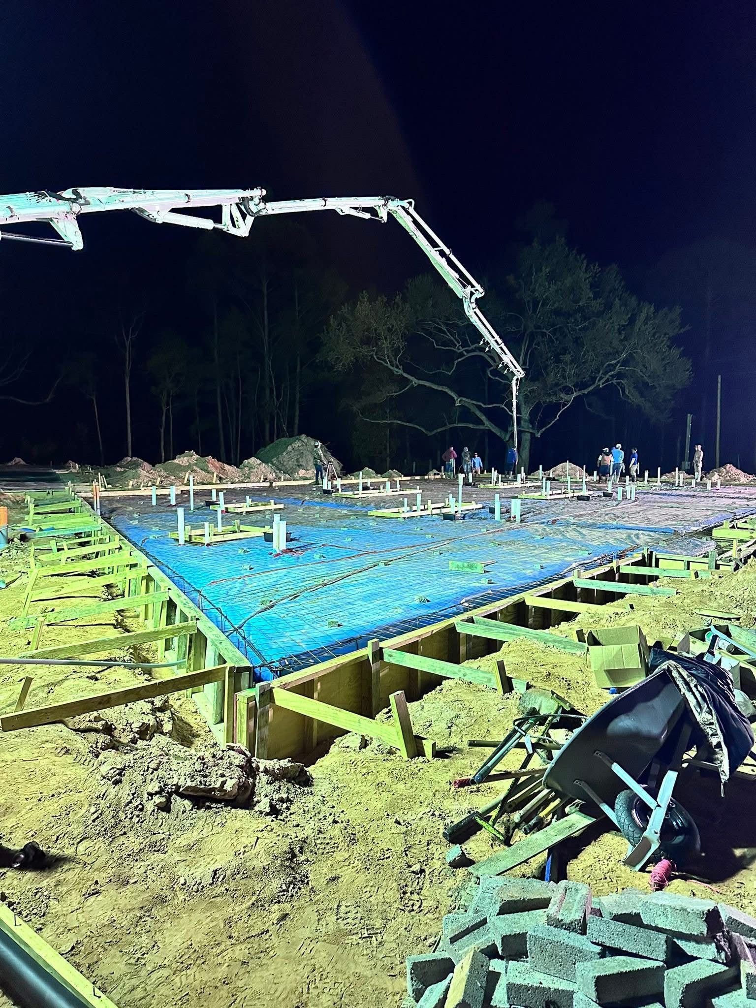 Nighttime construction site with a blue concrete slab, workers, and a crane arm overhead