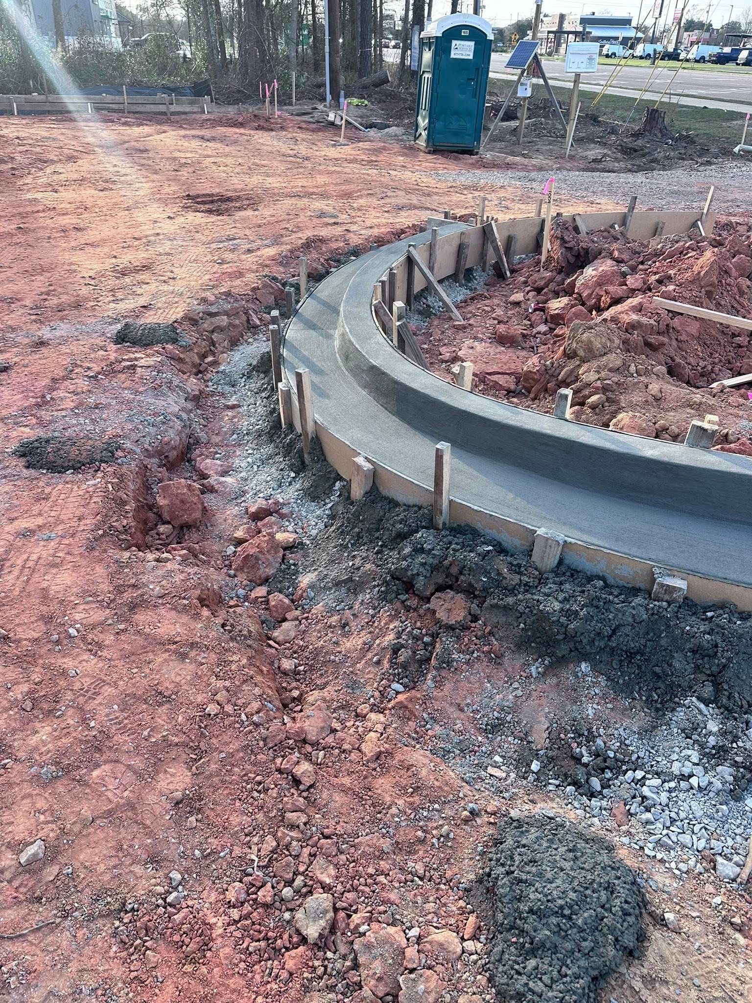 Curved concrete curb at a muddy construction site with a green portable toilet in the background