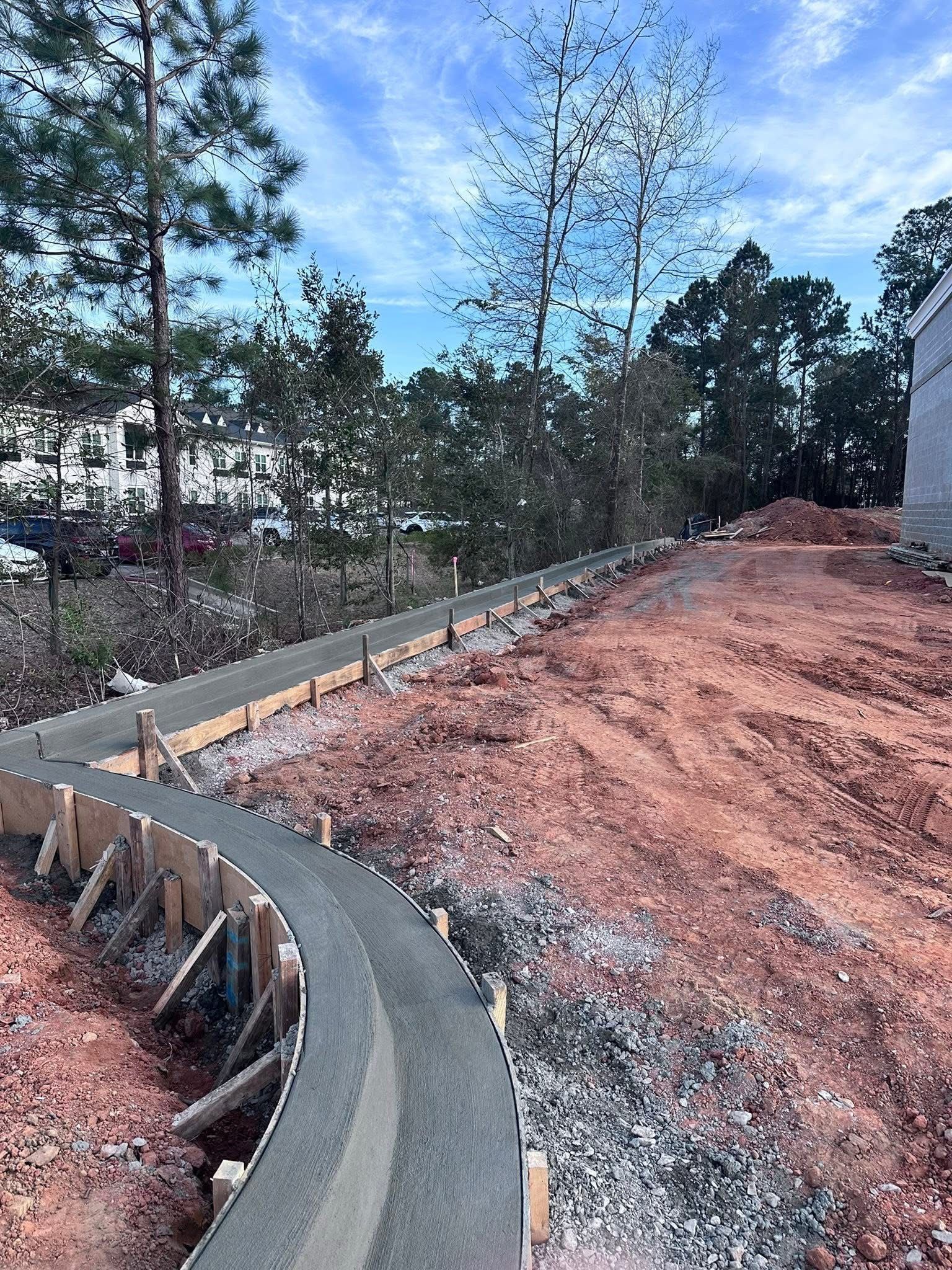 Curving concrete curb along a dirt roadside under construction, with trees and blue sky in the background