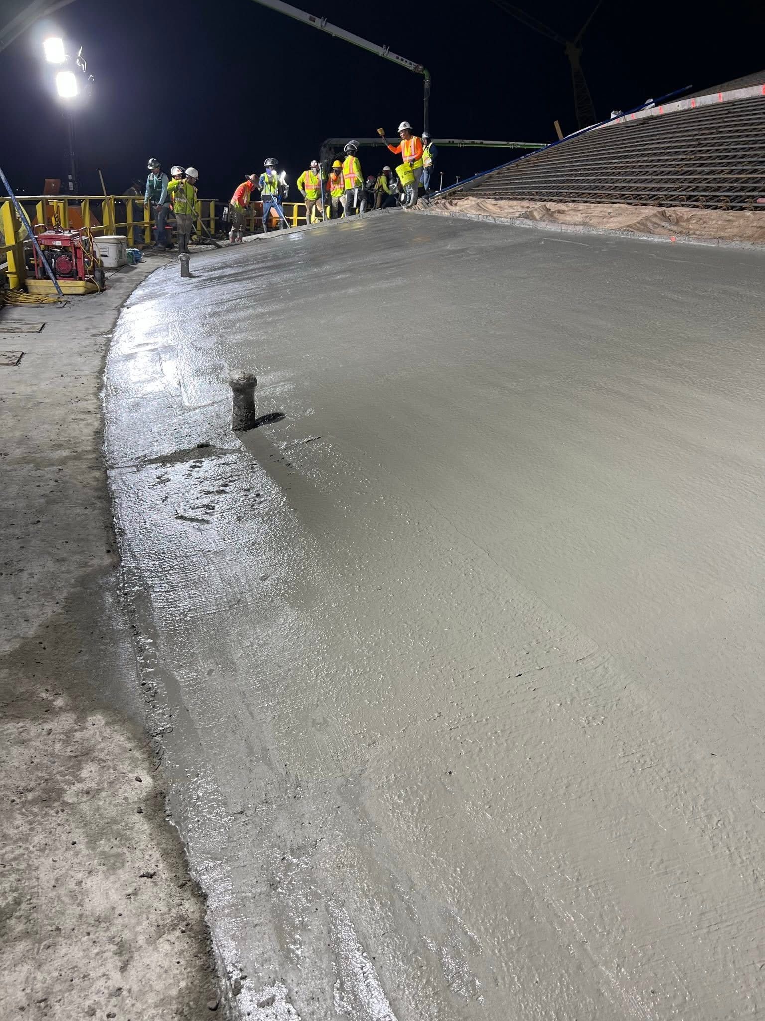 Night roadwork crew beside a freshly paved, wet road under bright floodlights at night
