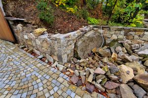 Stone retaining wall beside a cobblestone path, with scattered rocks and greenery on a slope
