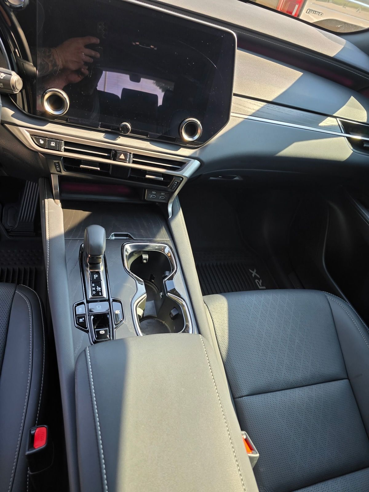 A front-seat interior view of a modern vehicle showing the center console, gear shifter, cup holders, and infotainment.