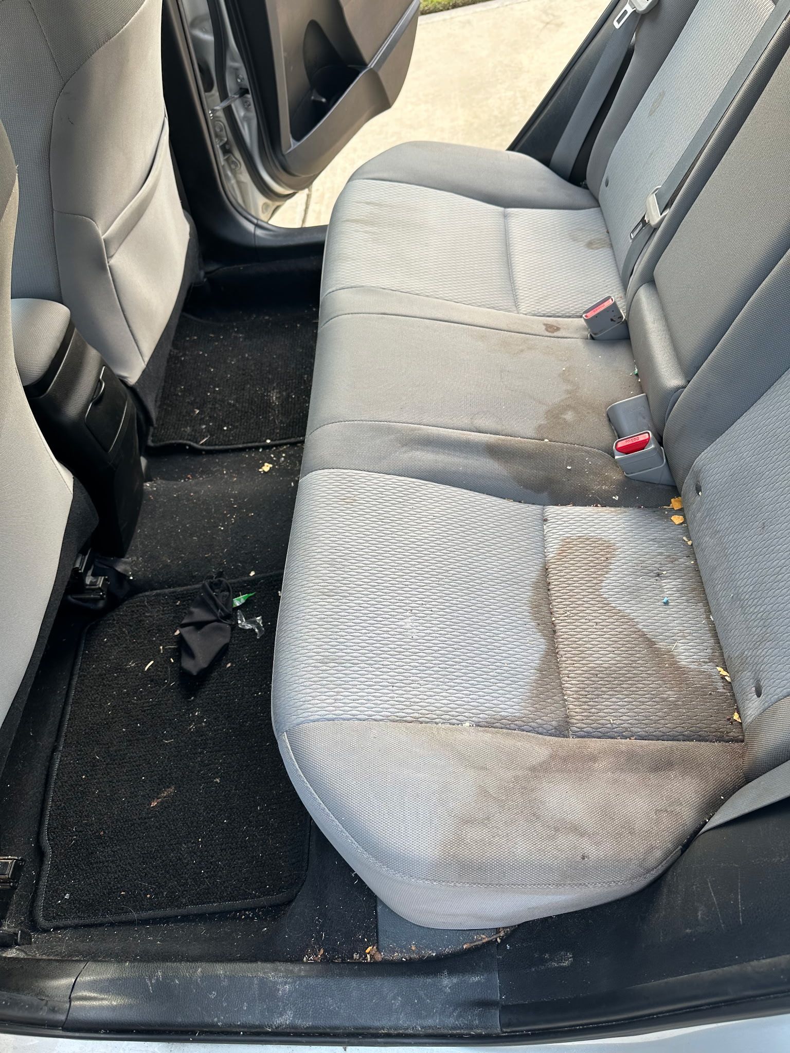 The interior backseat of a car showing grey fabric upholstery with noticeable stains and debris on the seats and floor.