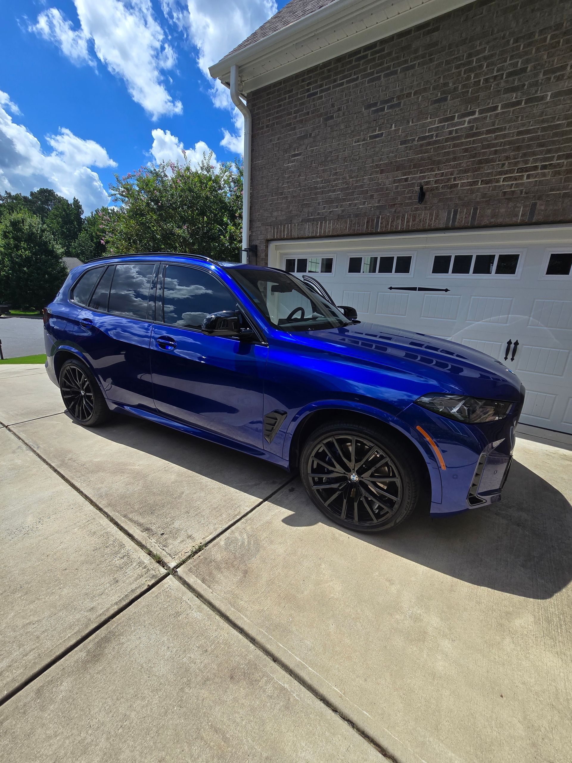 A blue BMW X5 SUV parked on a concrete driveway in front of a brick house garage under a sunny blue sky.