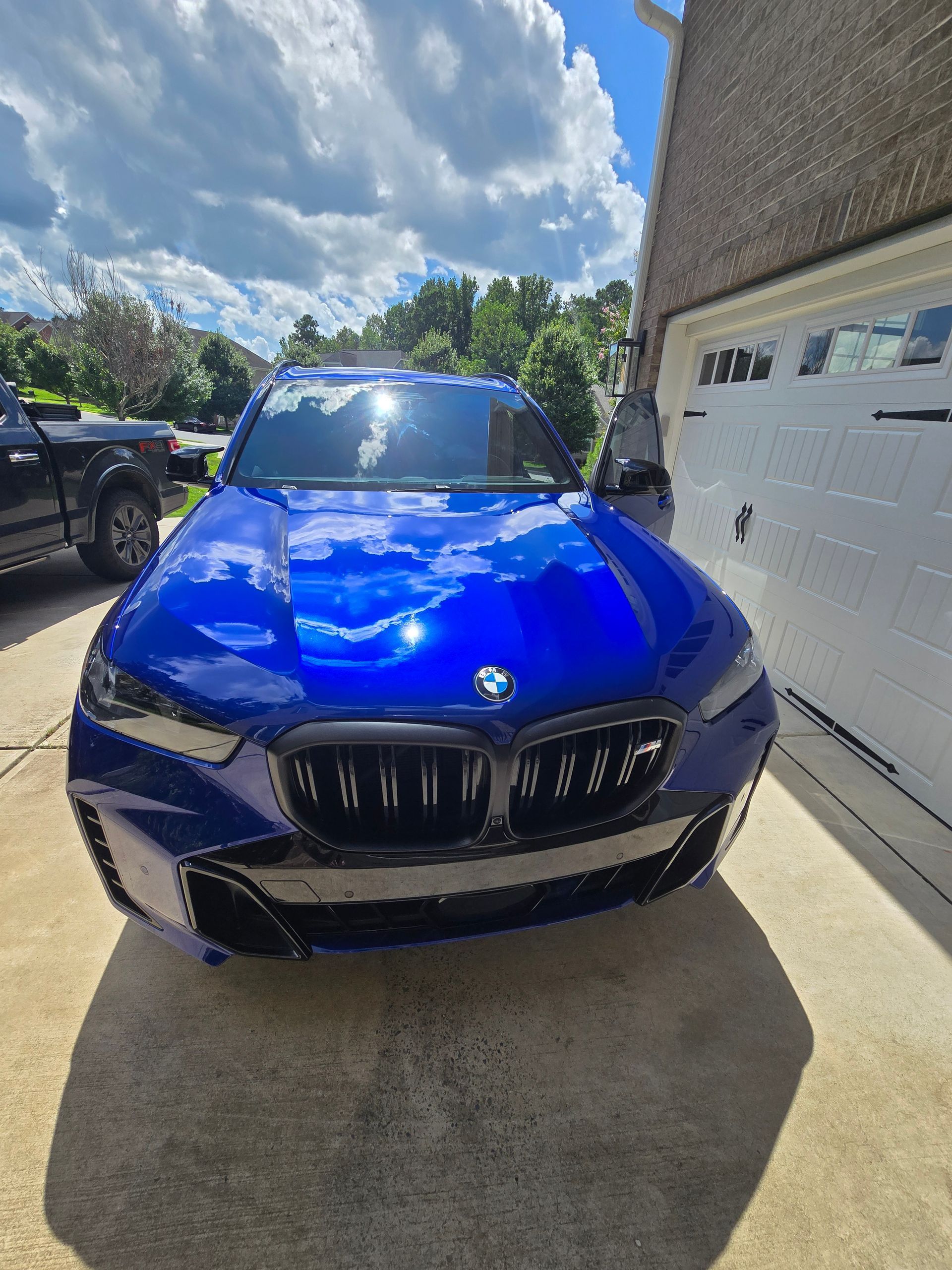 A bright blue BMW SUV parked on a residential driveway in front of a white garage door under a cloudy sky.