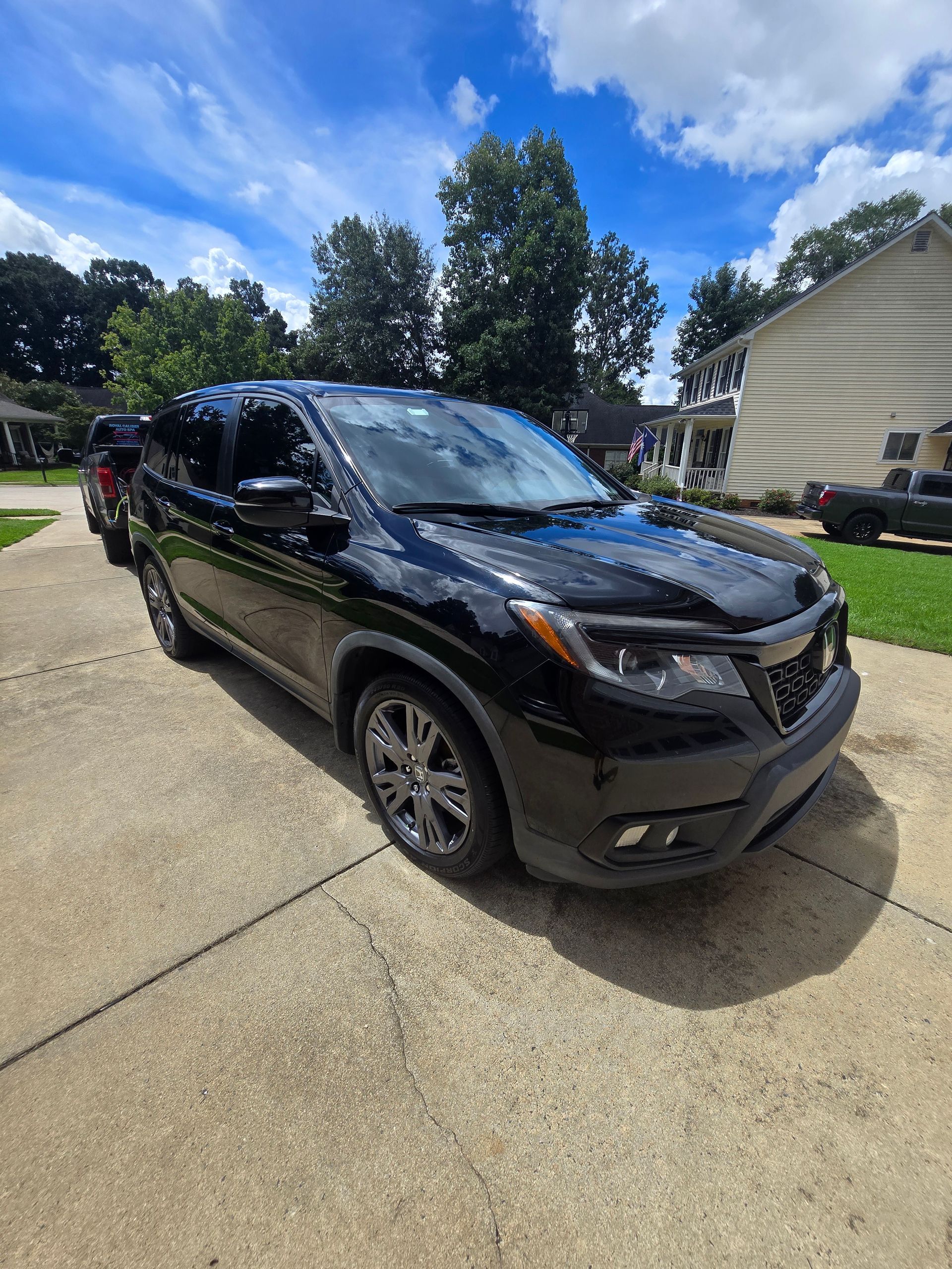 A black Honda Passport SUV parked on a residential concrete driveway under a blue, cloudy sky.