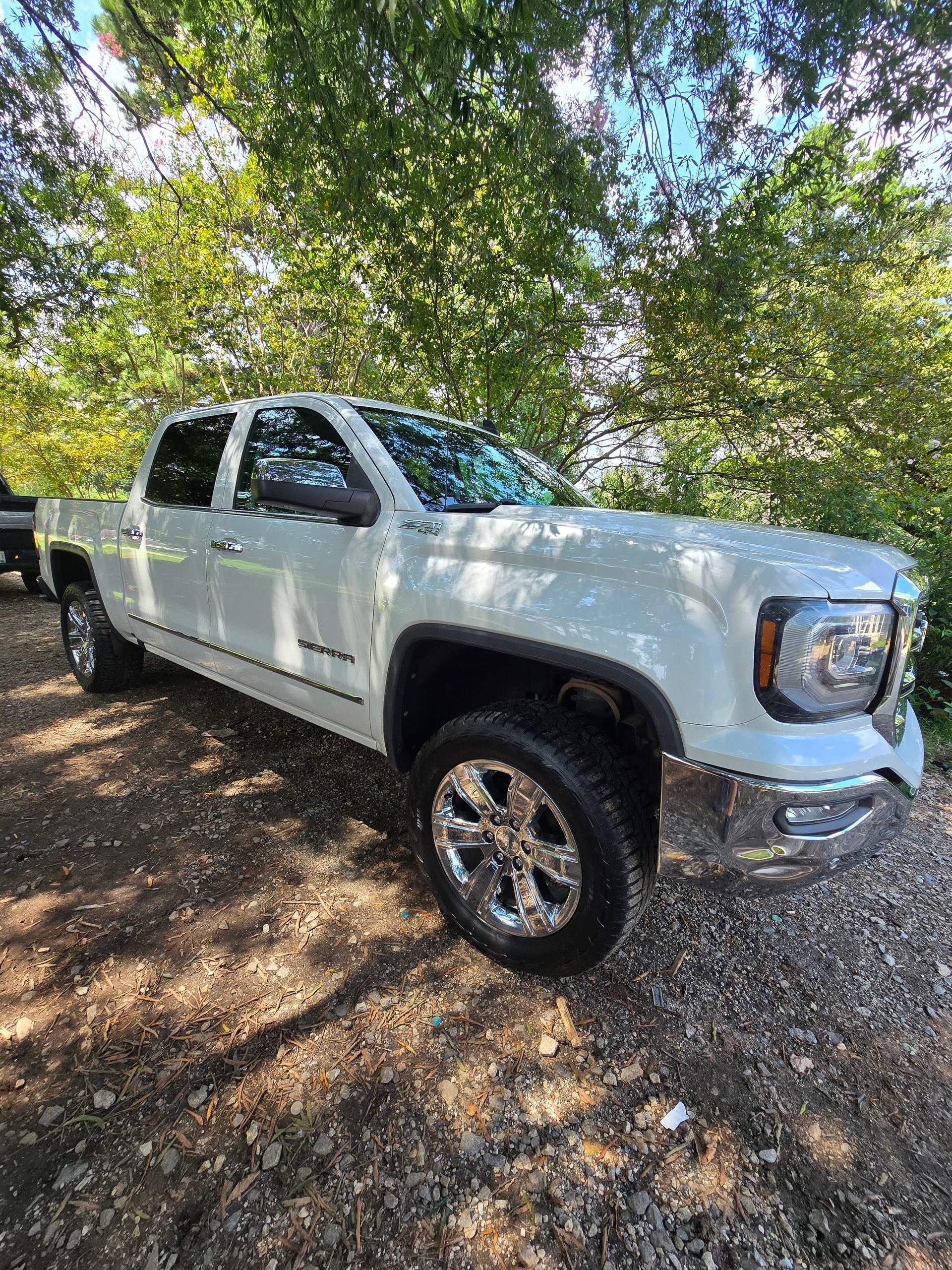 A white GMC Sierra crew cab pickup truck parked on a gravel path surrounded by trees.
