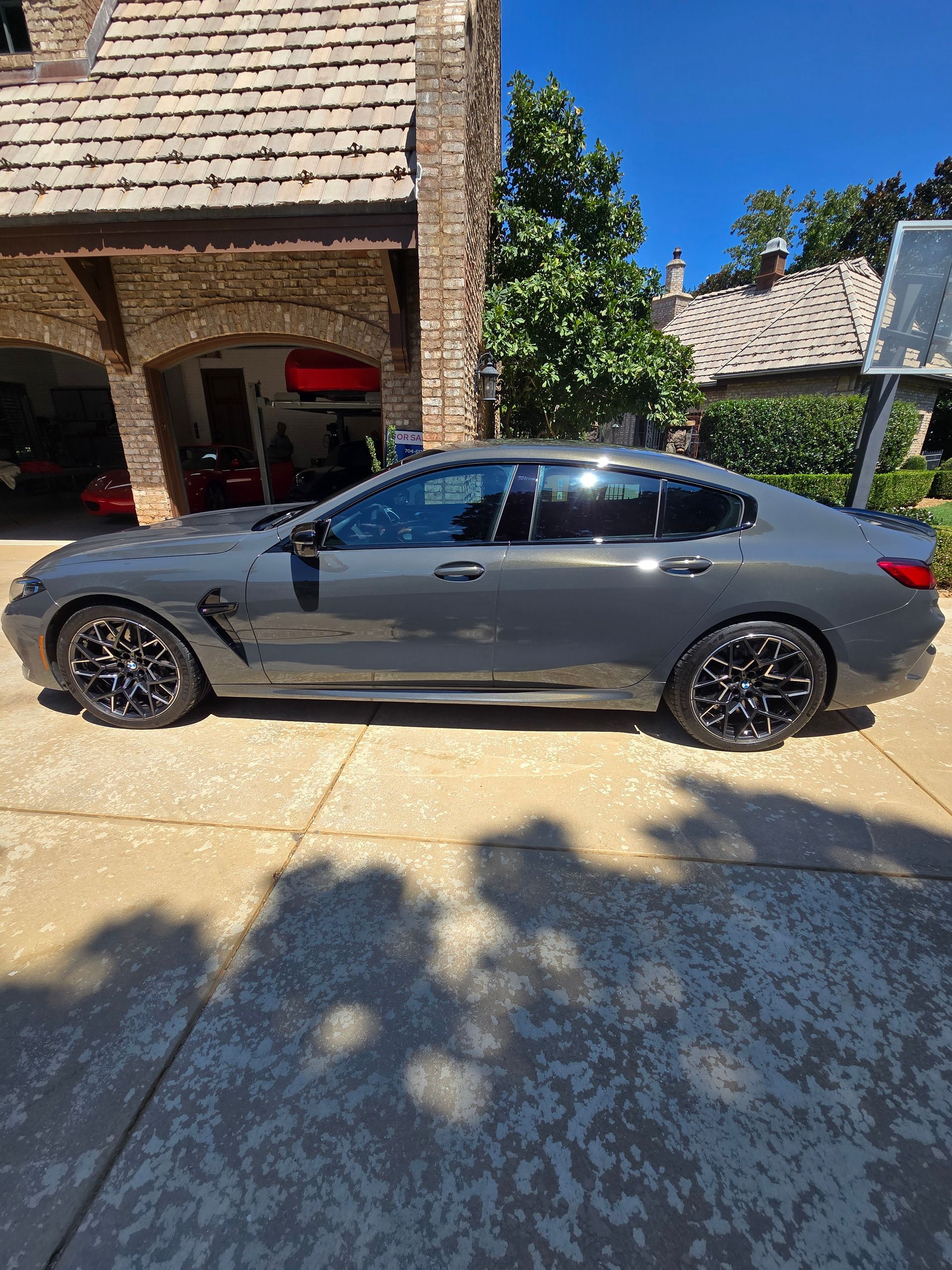 A dark gray BMW M3 sedan parked in a sunny driveway in front of a stone house with an arched garage.