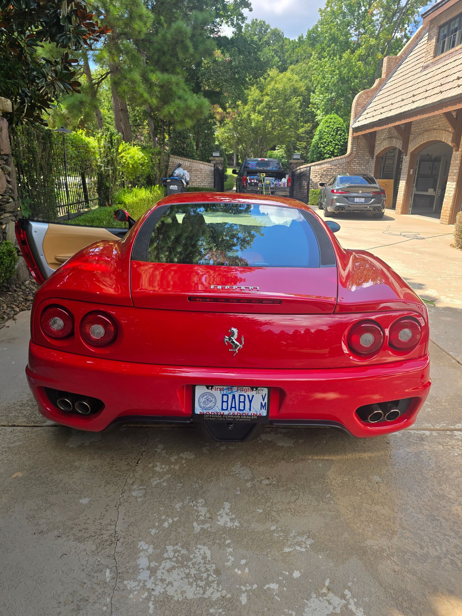 A red Ferrari parked on a stone driveway in front of a house, seen from the rear with its driver-side door open.