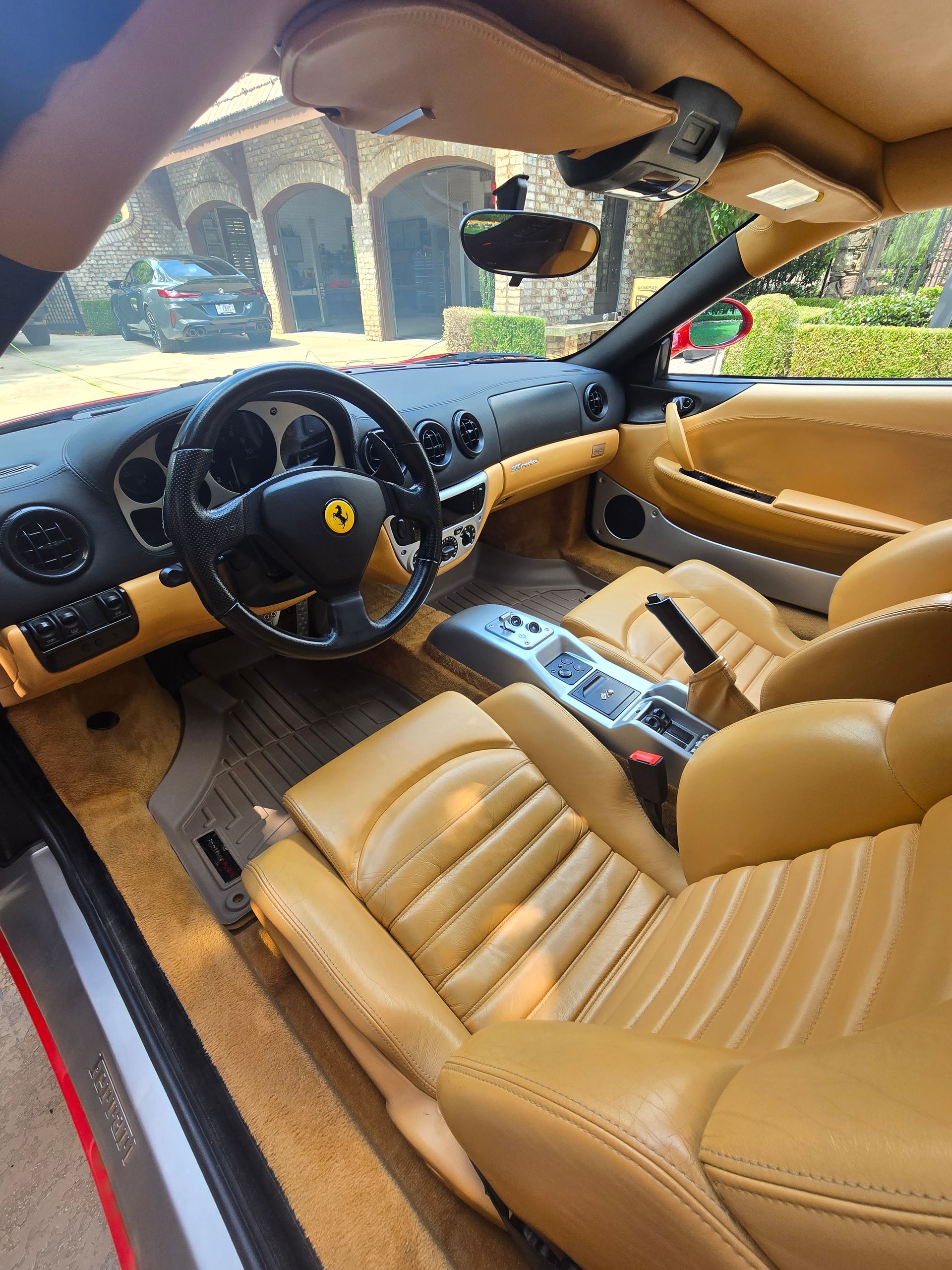 The interior of a Ferrari, featuring tan leather bucket seats, a black dashboard, and a black steering wheel with a logo.