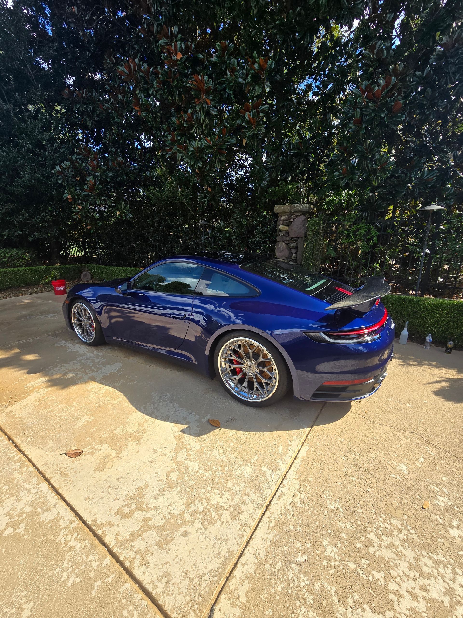 A dark blue Porsche 911 parked on a paved driveway in front of a dense green hedge.