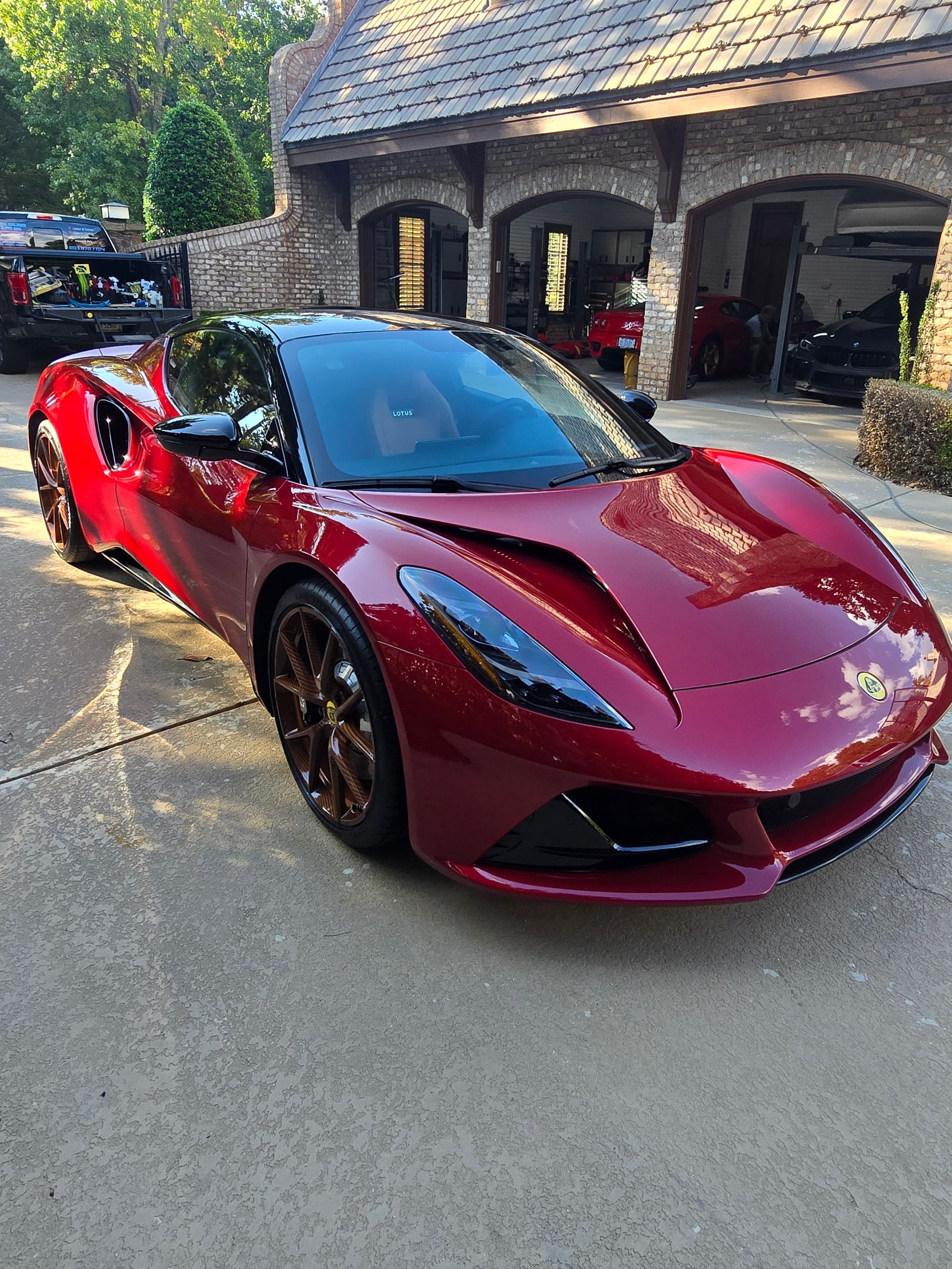 A deep red Lotus Emira sports car parked on a concrete driveway in front of a stone garage.