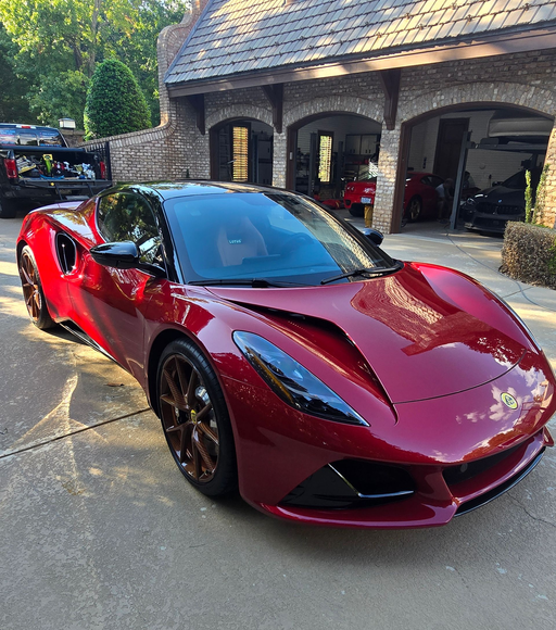A metallic red Lotus Emira sports car parked on a driveway in front of a stone garage.