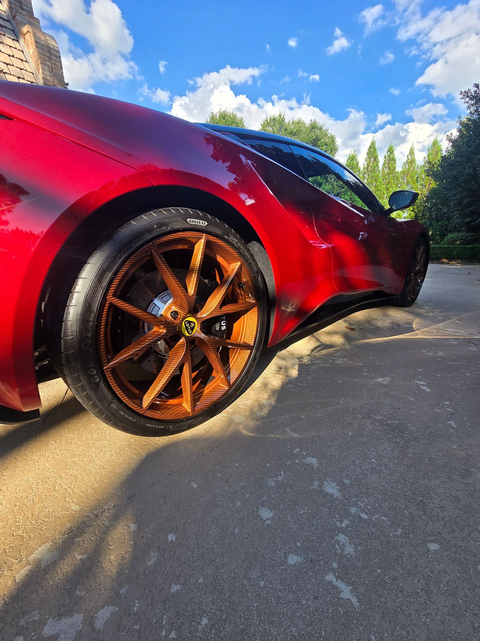 A red sports car with unique, rustic, dark-toned wheels parked on a paved surface under a bright blue sky.