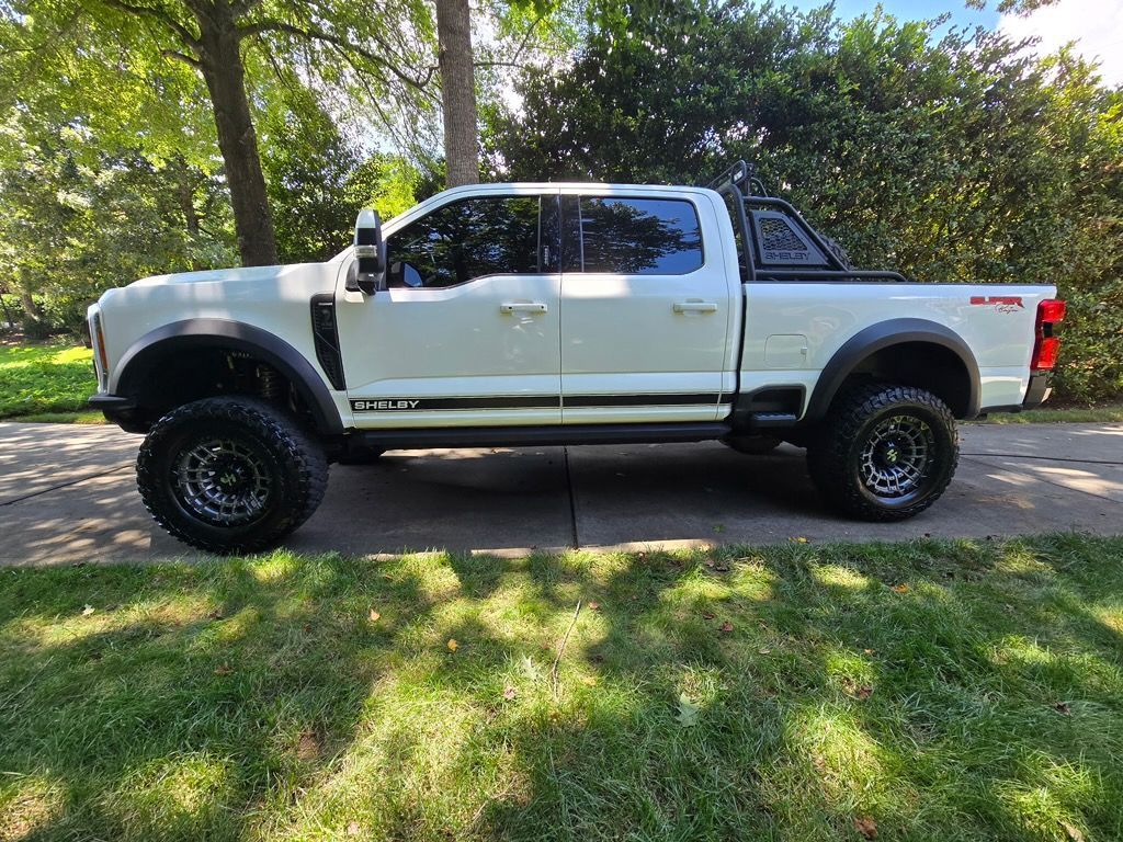 A white Shelby F-250 pickup truck with black off-road tires and a bed rack, parked on a driveway beside green trees.
