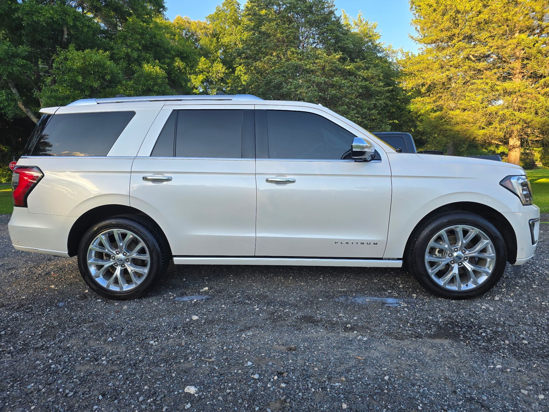 A white Ford Expedition Platinum SUV parked on a gravel surface outdoors with green trees in the background.