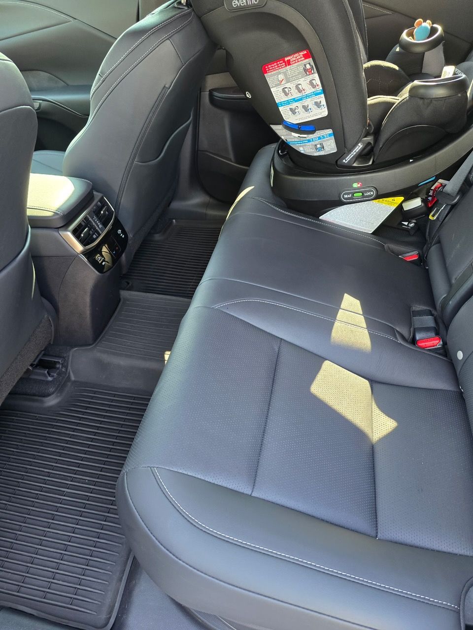 A child car seat installed on the rear black leather seat of a vehicle, with floor mats visible in the footwell.