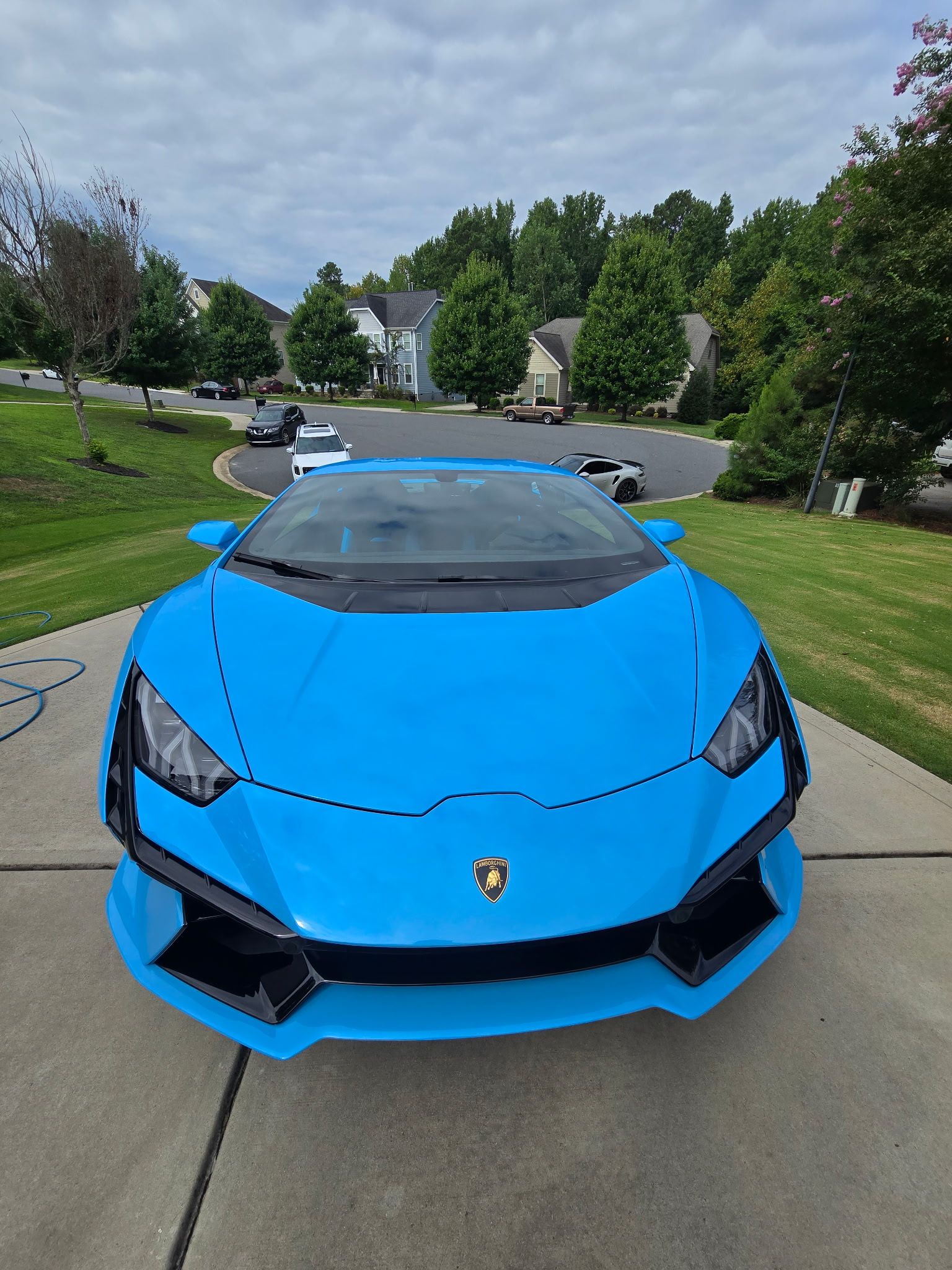 A bright blue Lamborghini sports car is parked on a suburban concrete driveway on a partly cloudy day.