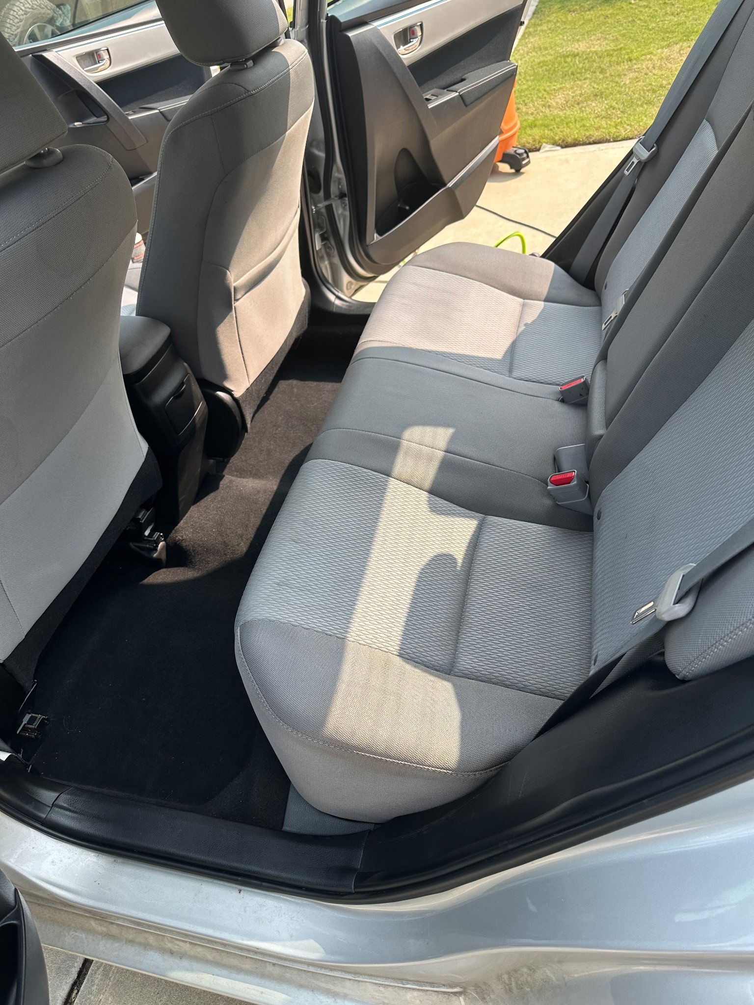 The back interior of a silver car, featuring gray upholstered seats, a clean floor mat, and an open side door.