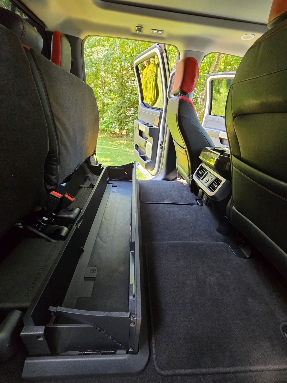 A black, under-seat storage bin installed in the back of a vehicle with a flat, carpeted floor and red-accented seats.