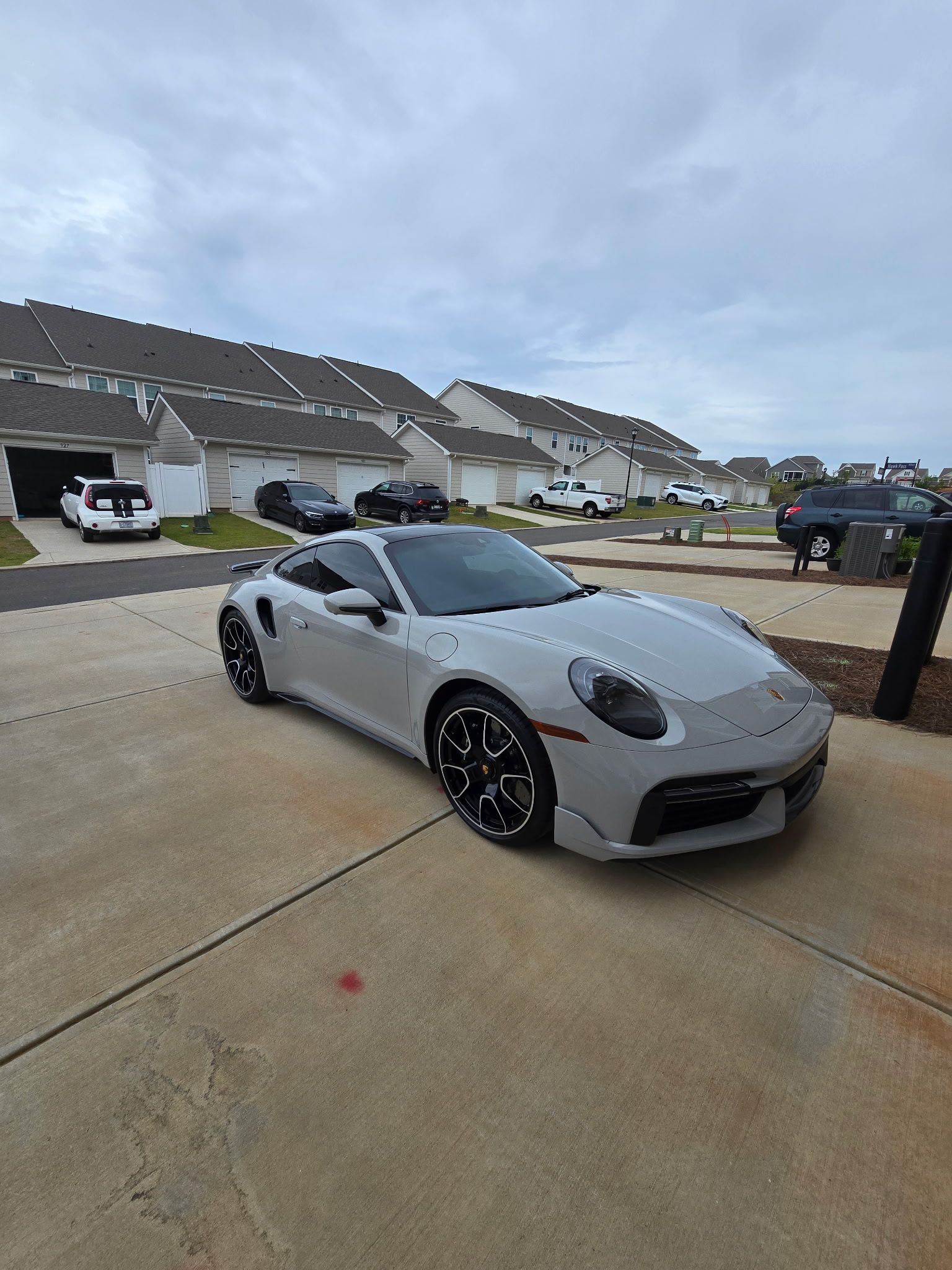 A light grey Porsche 911 sports car parked on a concrete driveway in front of a row of townhouses under a cloudy sky.