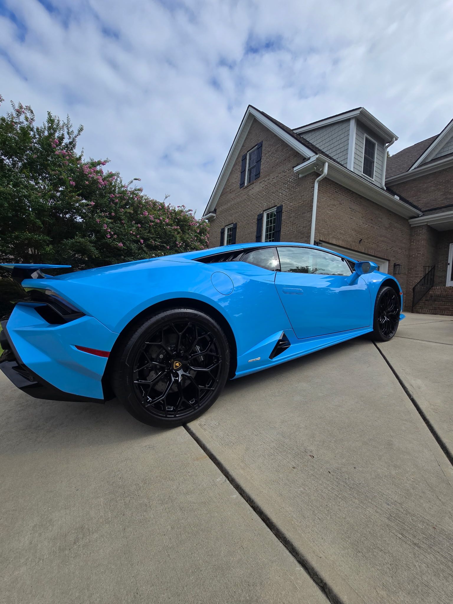 A bright blue Lamborghini Huracán parked on a concrete driveway in front of a brick house under a cloudy sky.