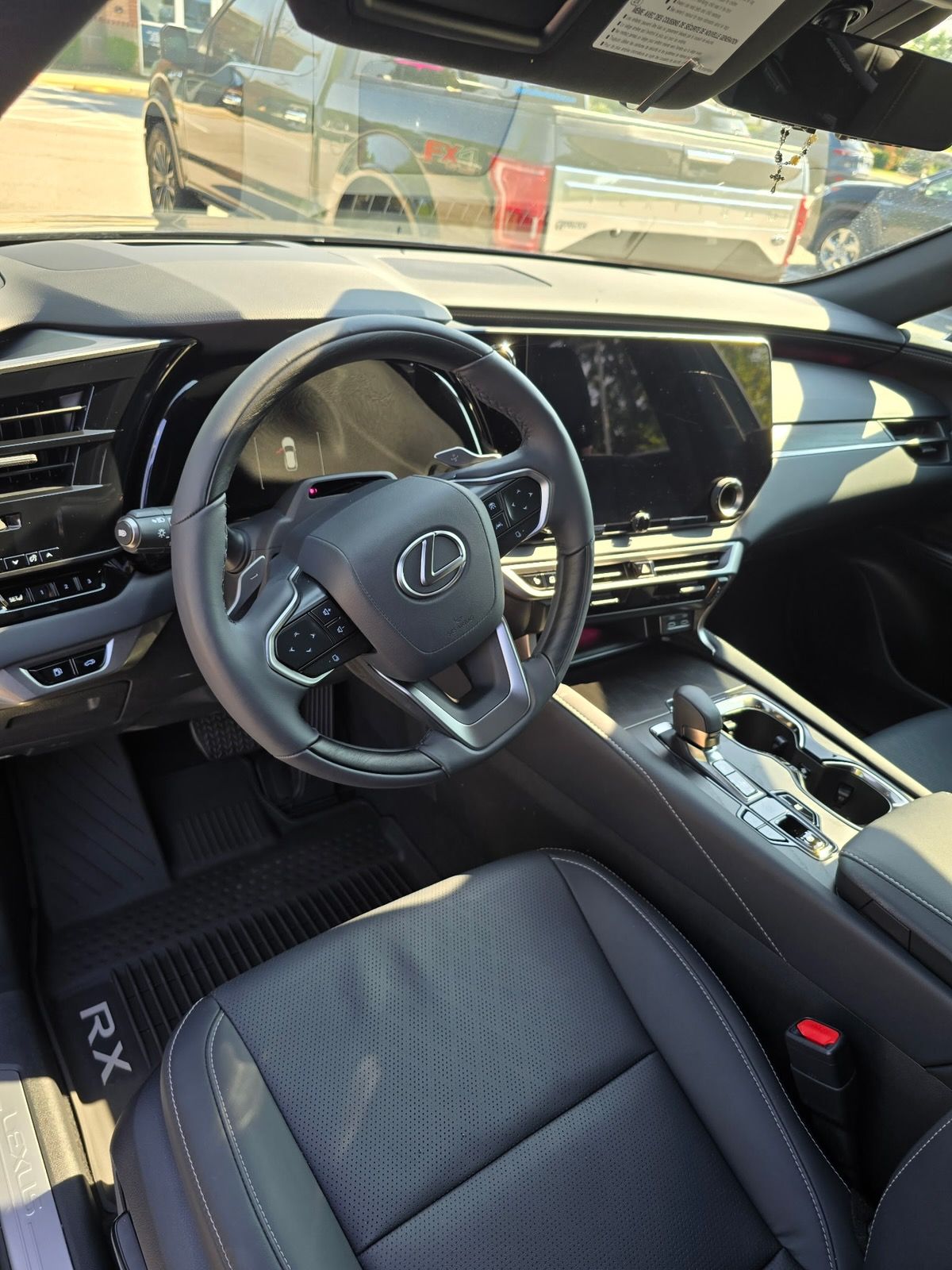 The interior of a Lexus RX, showing the steering wheel with the brand logo, dashboard screen, and dark leather front seat.