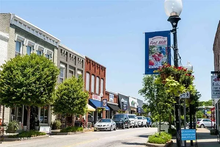 A street view of a small town with historic brick and stone buildings, leafy trees, and a banner hanging from a lamppost.