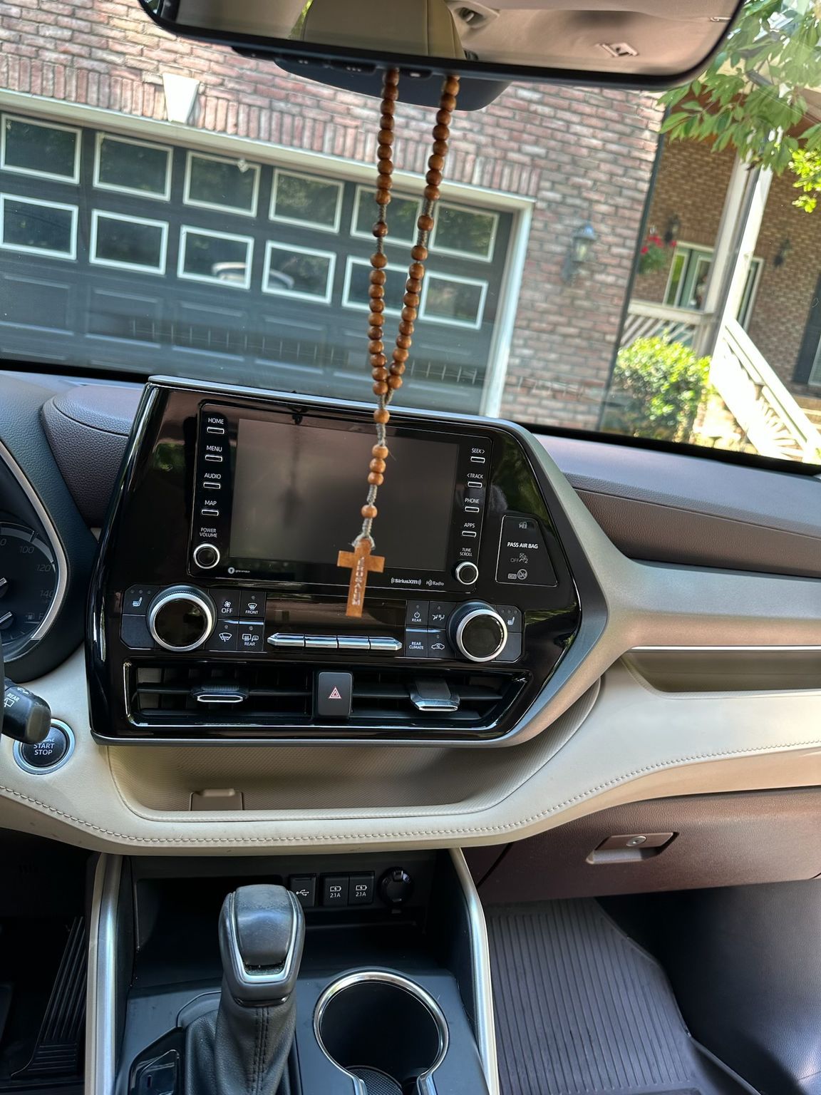 Interior of a car dashboard with a wooden rosary hanging from the rearview mirror against a house and driveway background.