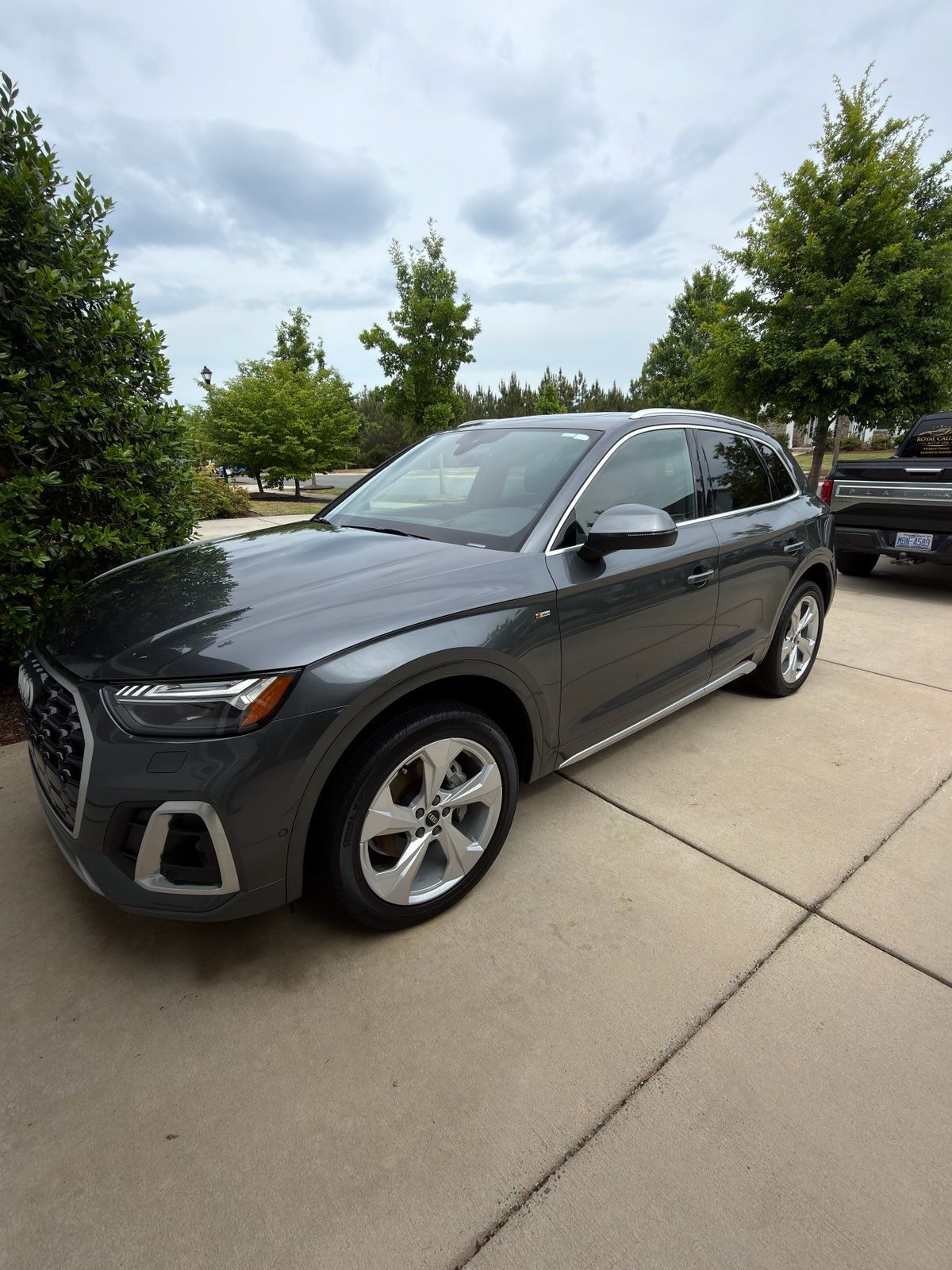 Gray SUV parked in a driveway beside trees and other cars under a cloudy sky