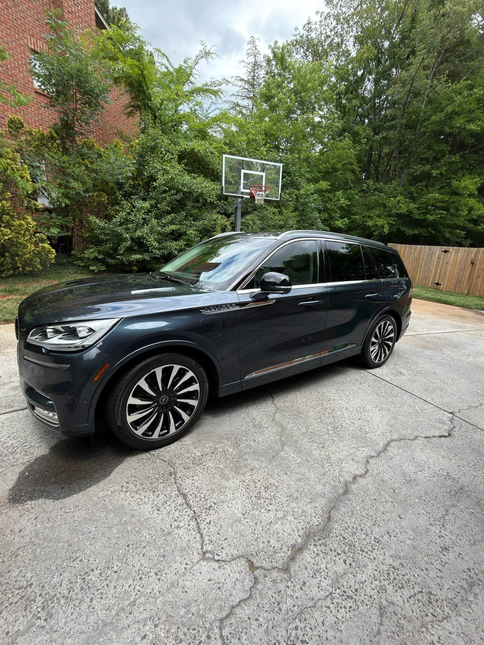 Dark SUV parked on a cracked driveway beside a basketball hoop and greenery