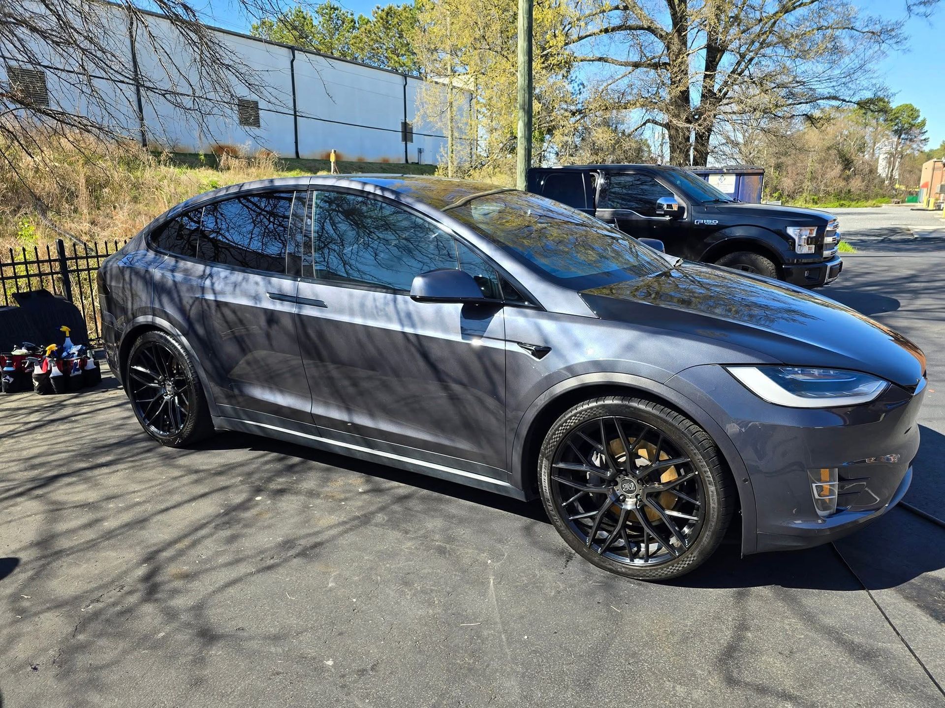 A dark gray Tesla Model X SUV with black custom wheels parked on an asphalt lot on a sunny day.