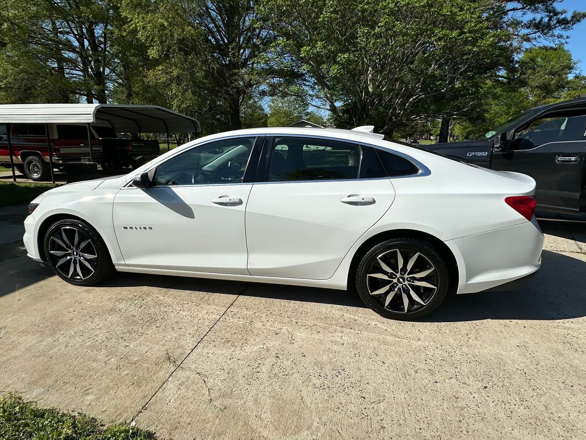 A white Chevrolet Malibu sedan parked on a concrete driveway in front of a shaded area with trees.