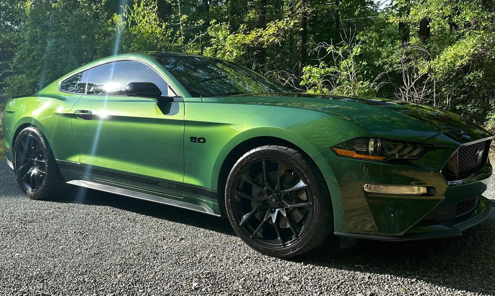 A metallic green Ford Mustang parked on a gravel surface with trees in the background.