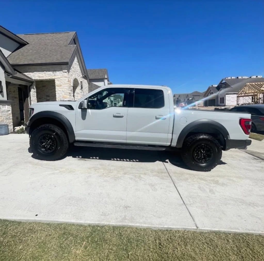 A light gray Ford F-150 Raptor pickup truck parked on a residential concrete driveway on a sunny day.