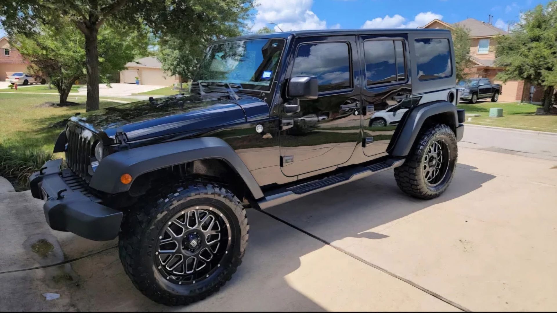A black Jeep Wrangler with custom wheels parked on a suburban driveway under a sunny sky.