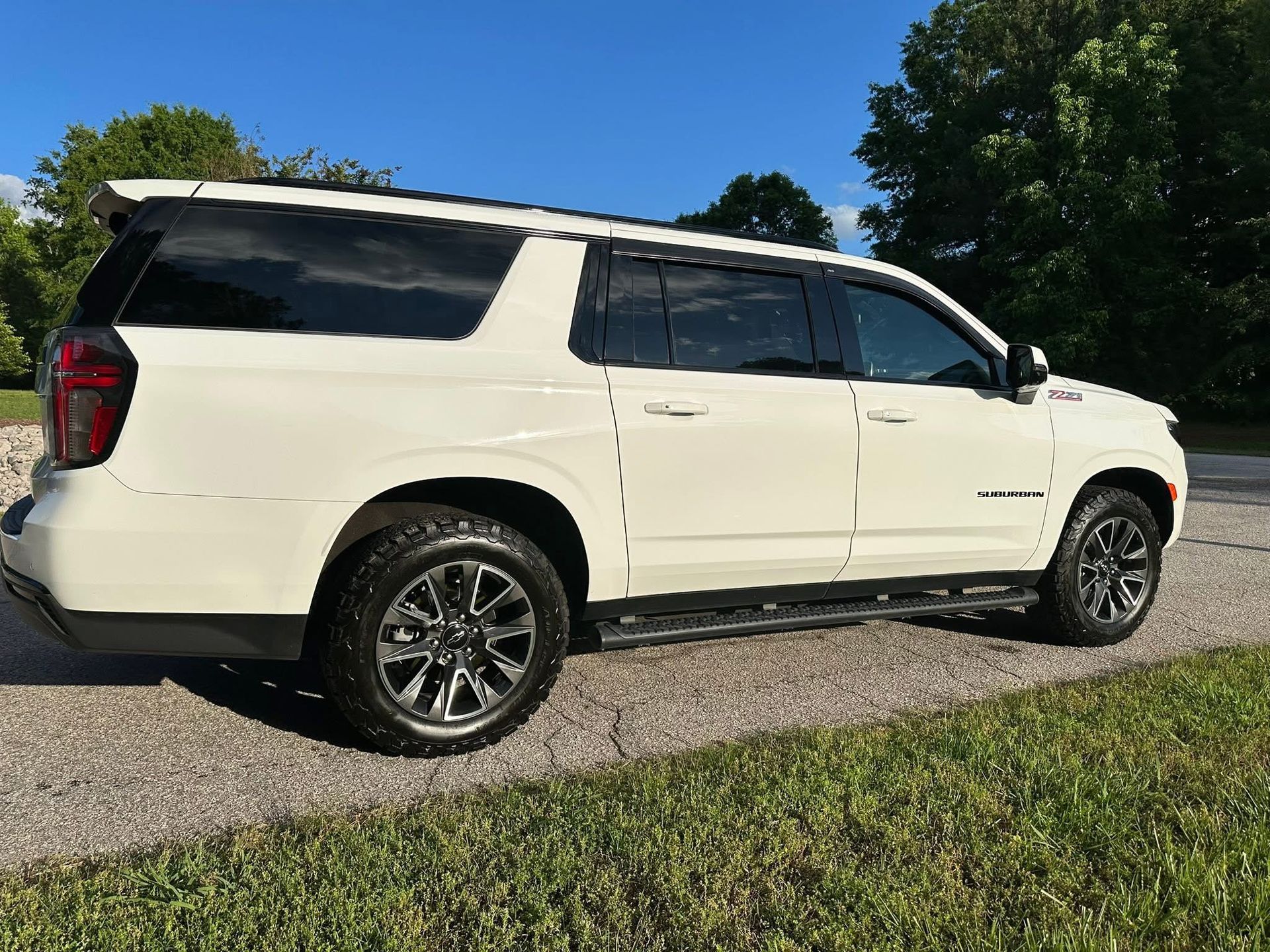 A side view of a white, extended-length SUV parked on a gravel surface against a blue sky and trees.