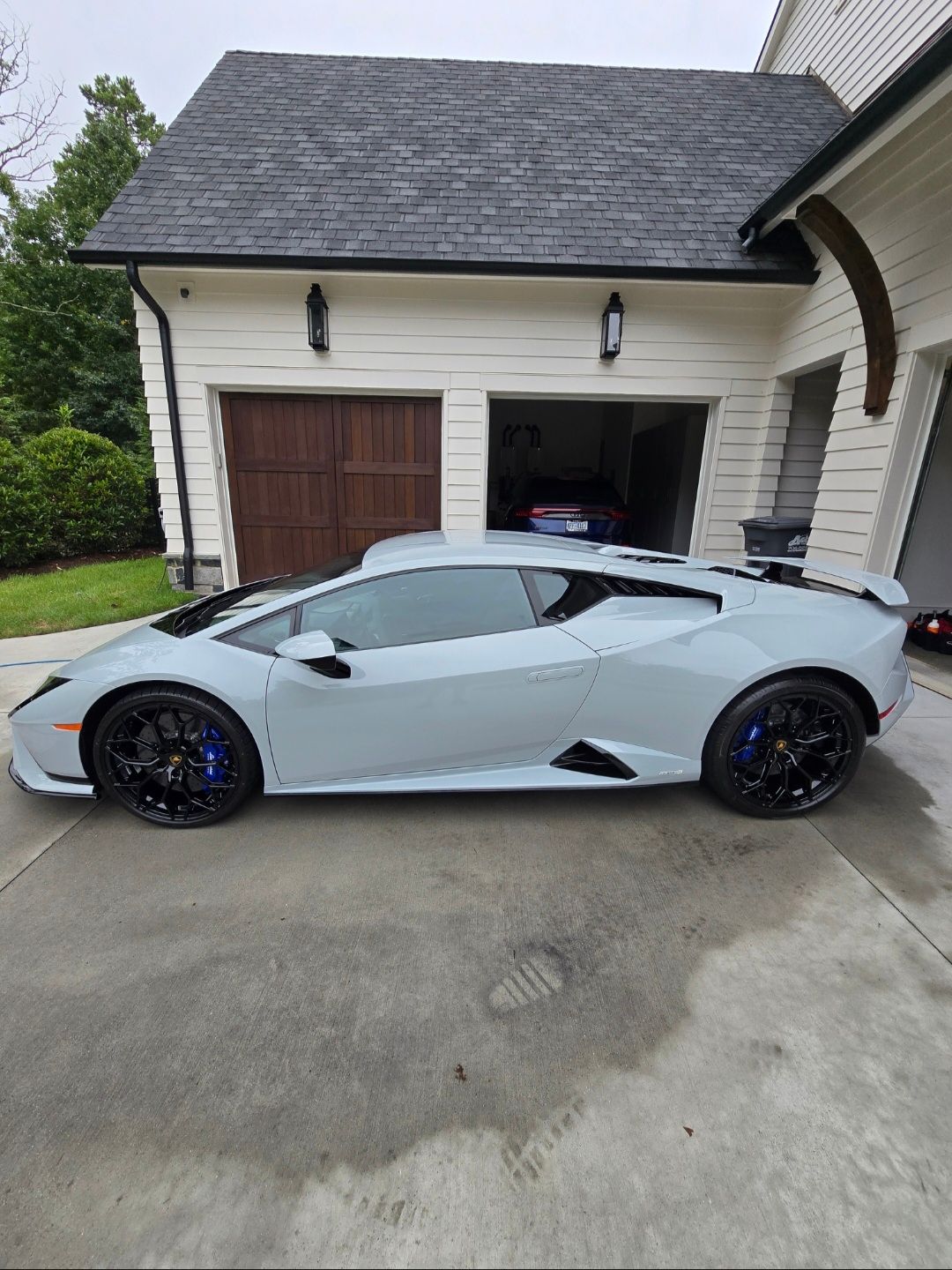 A light grey Lamborghini Huracán parked in the driveway in front of a house with a multi-car garage.